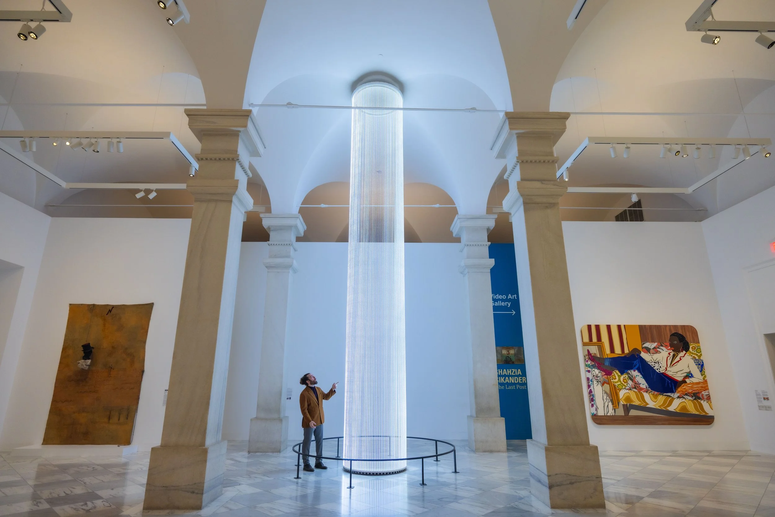 Interior view of an art gallery with white walls and arched ceiling. A large digital column artwork is in the center, with a man pointing at it and another person looking up. Artwork pieces are on the walls, and a blue sign with white text reads "Vid