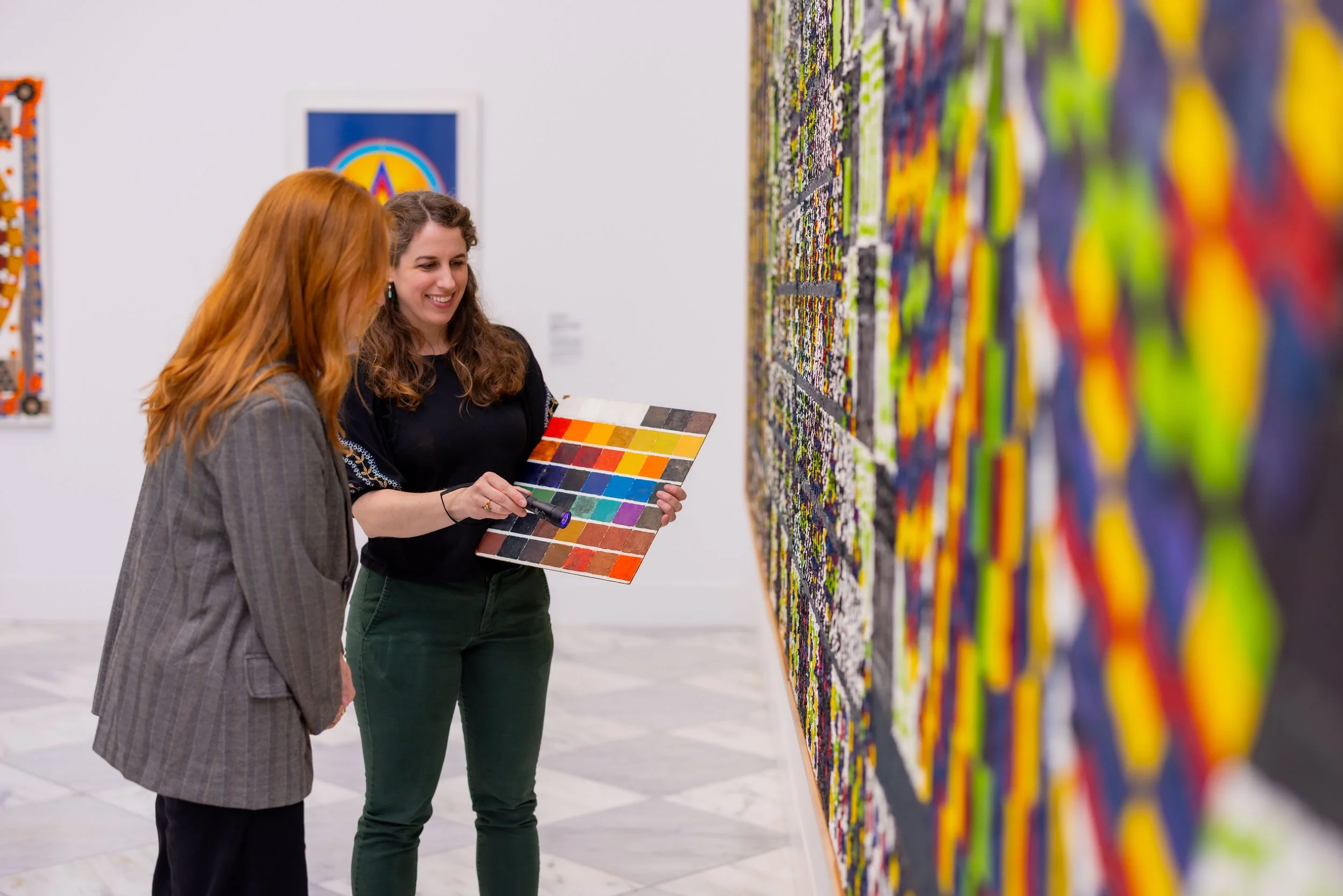 Two women look at colorful abstract artwork in an art gallery, one woman holds a color palette and a flashlight, smiling while viewing the art.