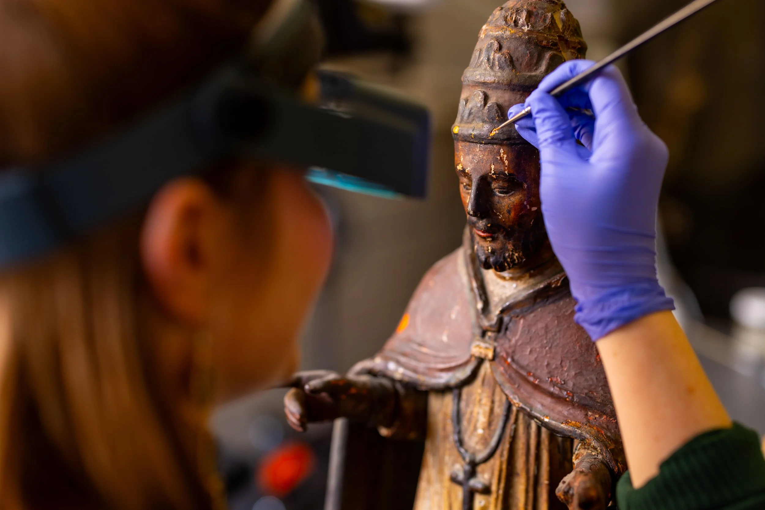 Close-up of a person wearing blue gloves carefully restoring an ancient wooden sculpture.