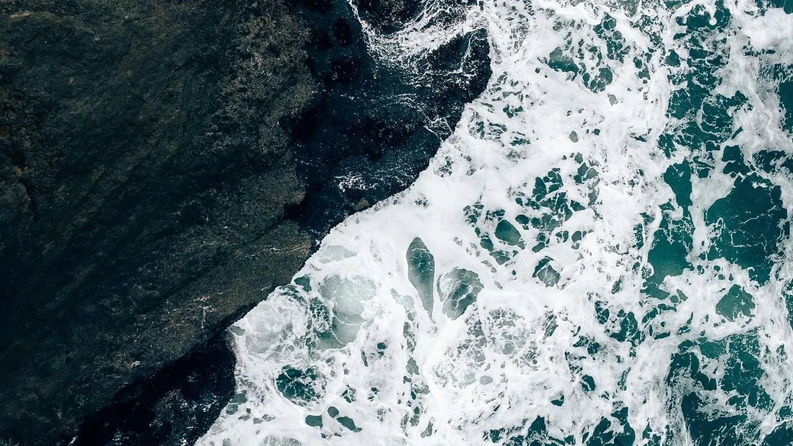 Aerial view of white surf breaking against dark coastal rocks