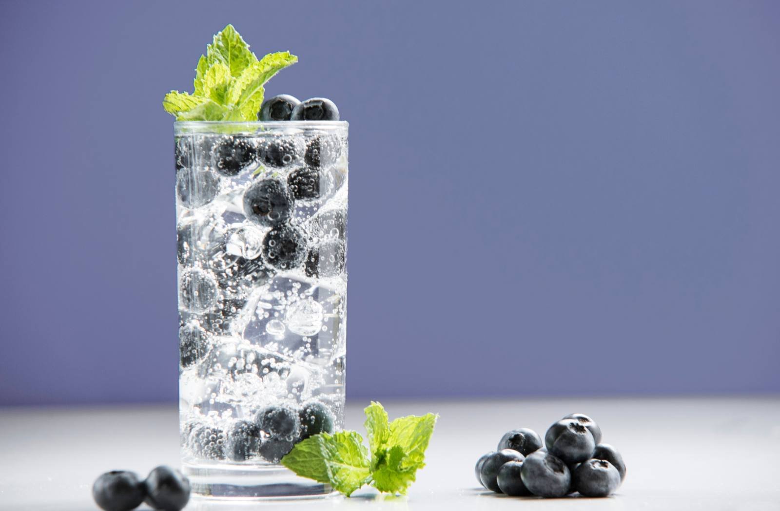 Tall glass of sparkling water with blueberries and mint on purple background