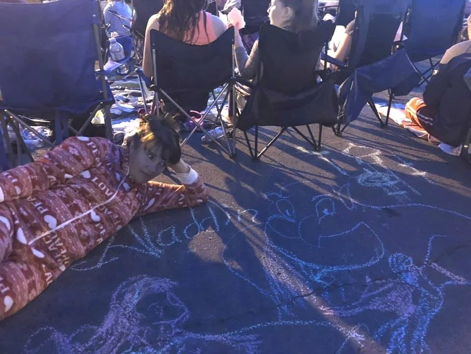 Person in red onesie lying on the ground with chalk drawings on pavement, surrounded by chairs and people in an outdoor setting.