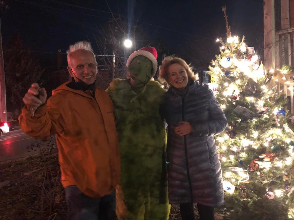 Two people posing with a person dressed as the Grinch in front of a decorated Christmas tree with lights at night.