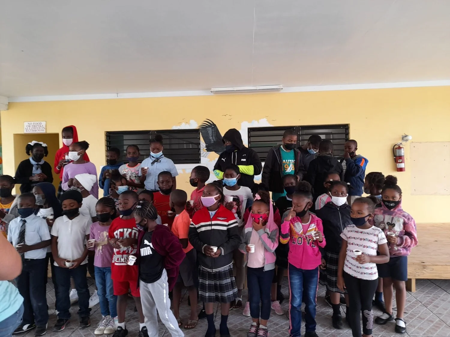 Group of children wearing masks indoors, holding cups, standing in front of a yellow wall.