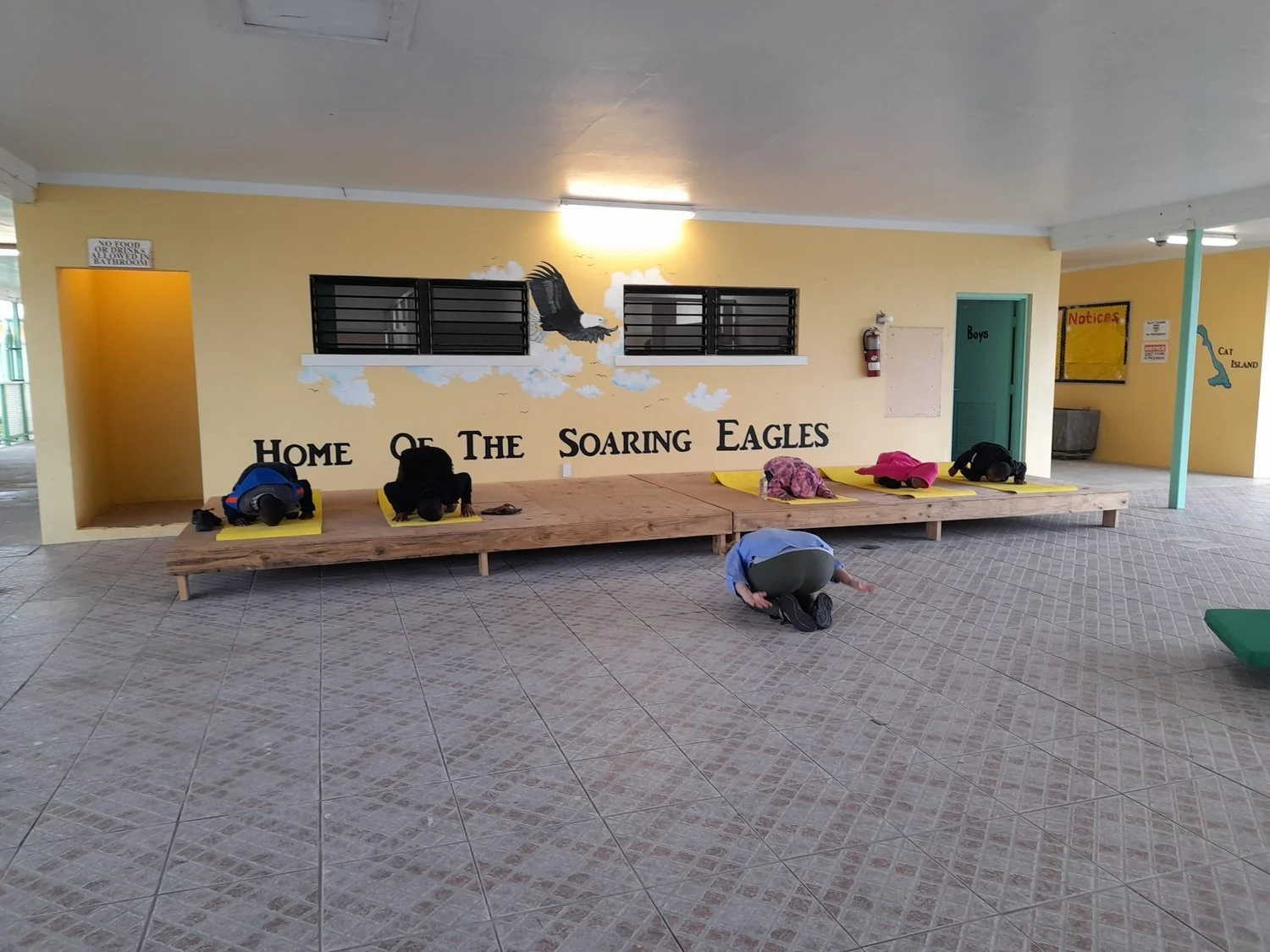 Children practicing yoga in child's pose on yellow mats in a school hallway with "Home of The Soaring Eagles" painted on the wall.
