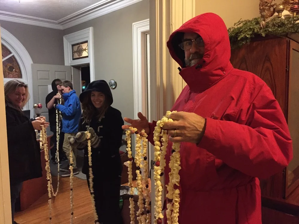 Group of people indoors holding handmade popcorn garlands, wearing winter clothing.