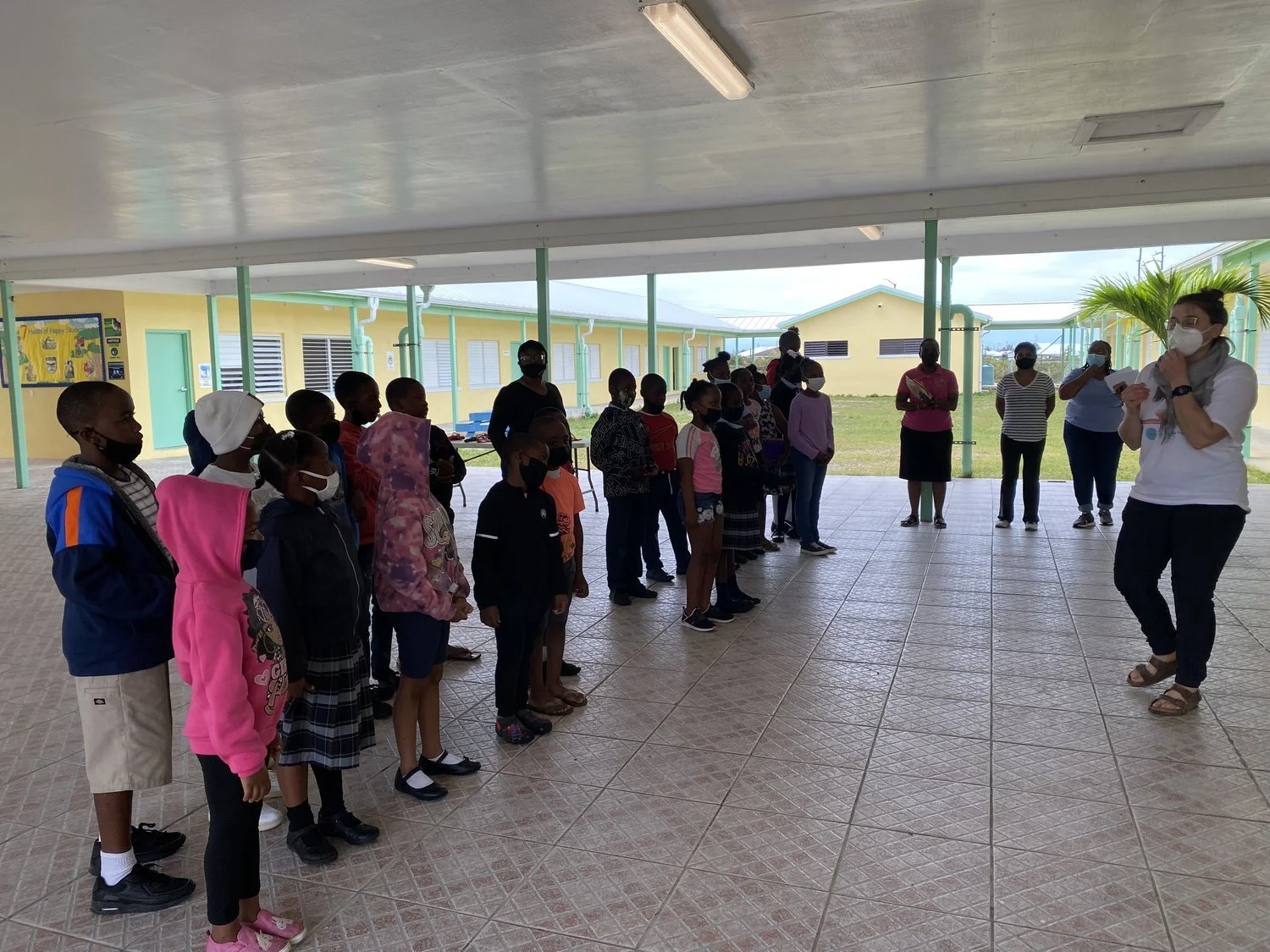 A group of children in masks stand in a line, facing an adult instructor also wearing a mask. They are in a covered outdoor area with yellow buildings in the background. Several other adults are observing the scene.