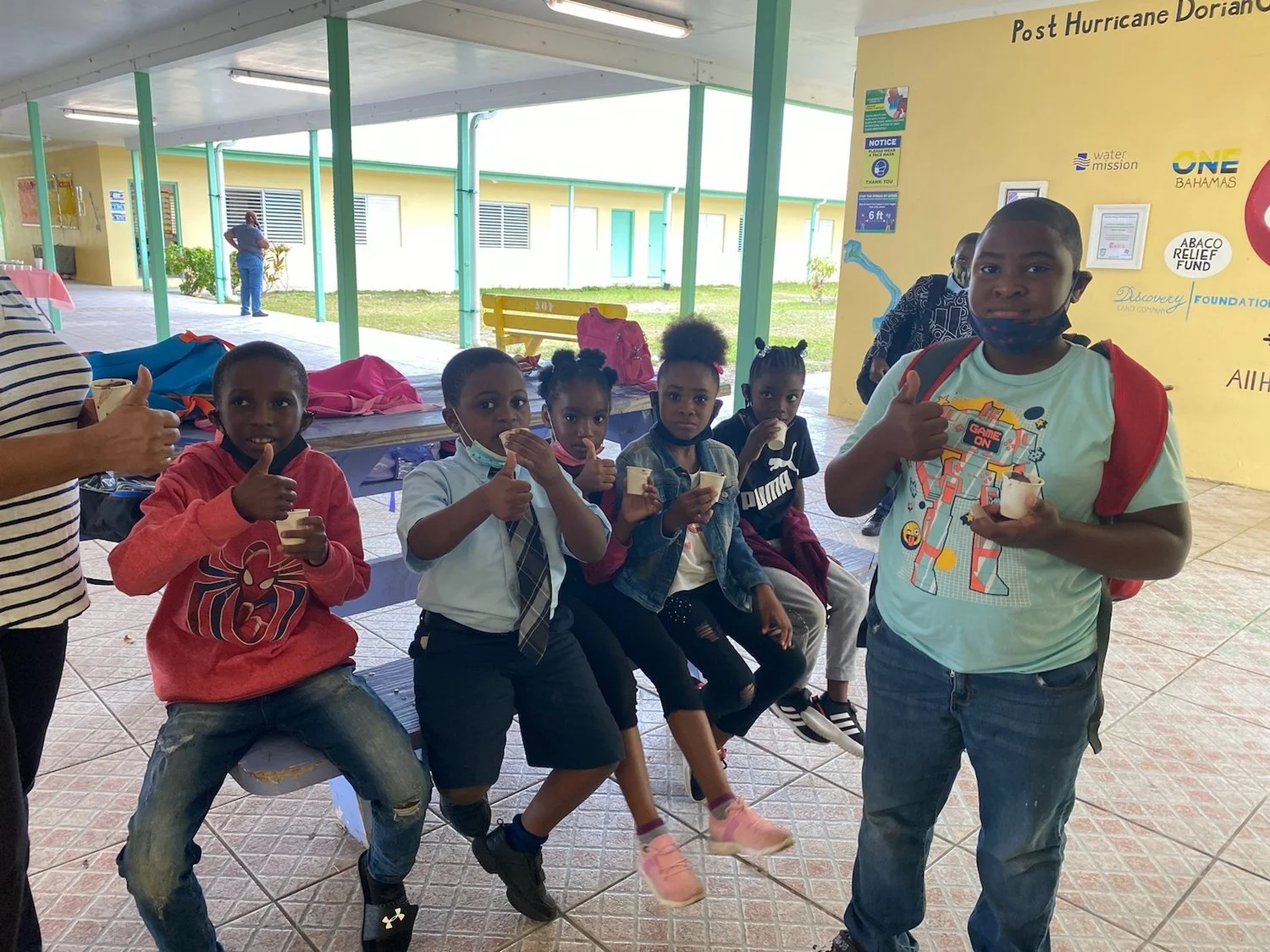 Children sitting on a bench, indoors, with colorful clothing, holding cups, and showing thumbs up. Background shows a yellow wall with various posters and logos.