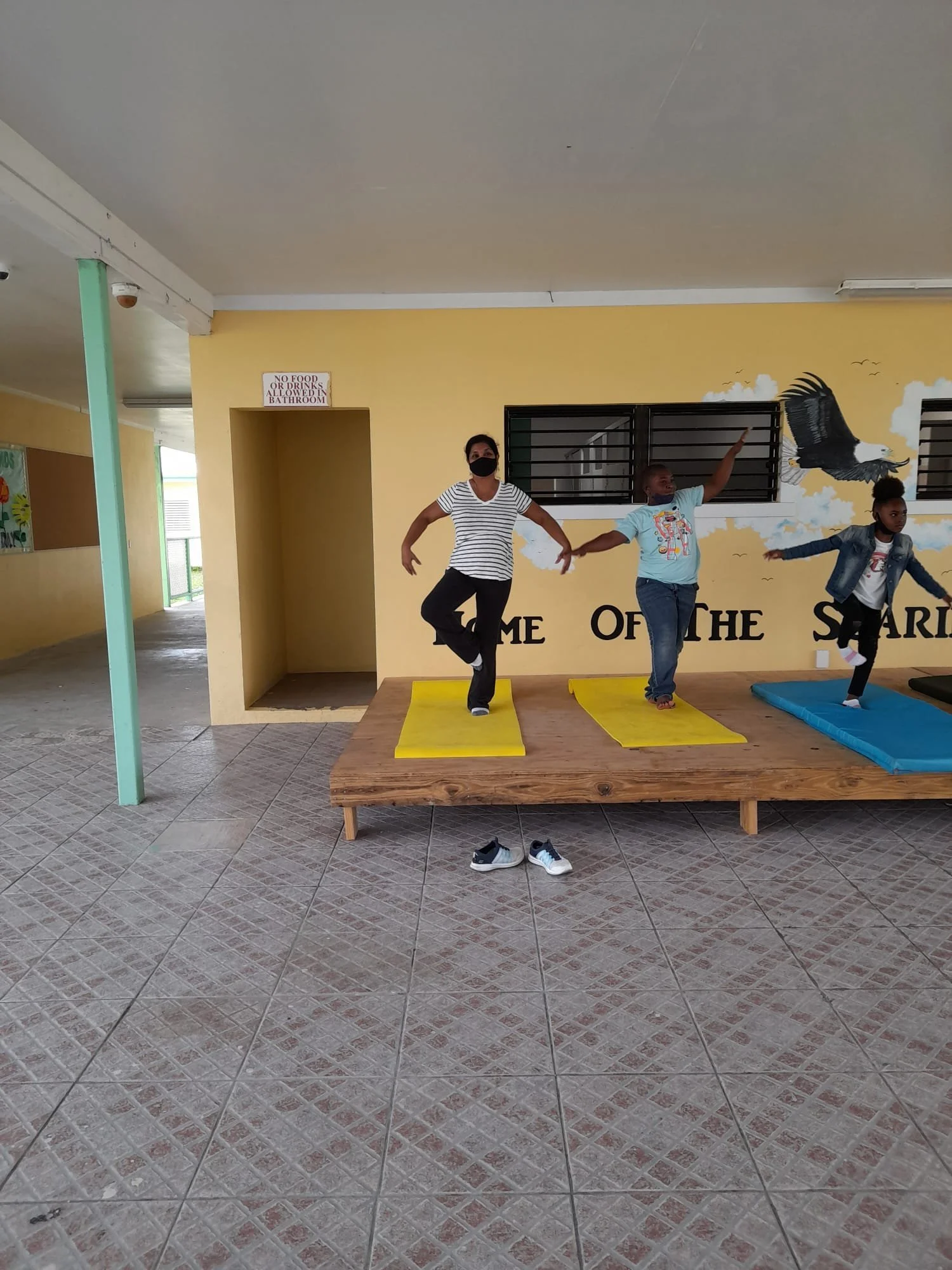 Three people balancing on one leg on mats indoors, with two yellow mats and one blue. Behind them is a mural with an eagle and partially visible text. Shoes are on the floor in front of the mats.