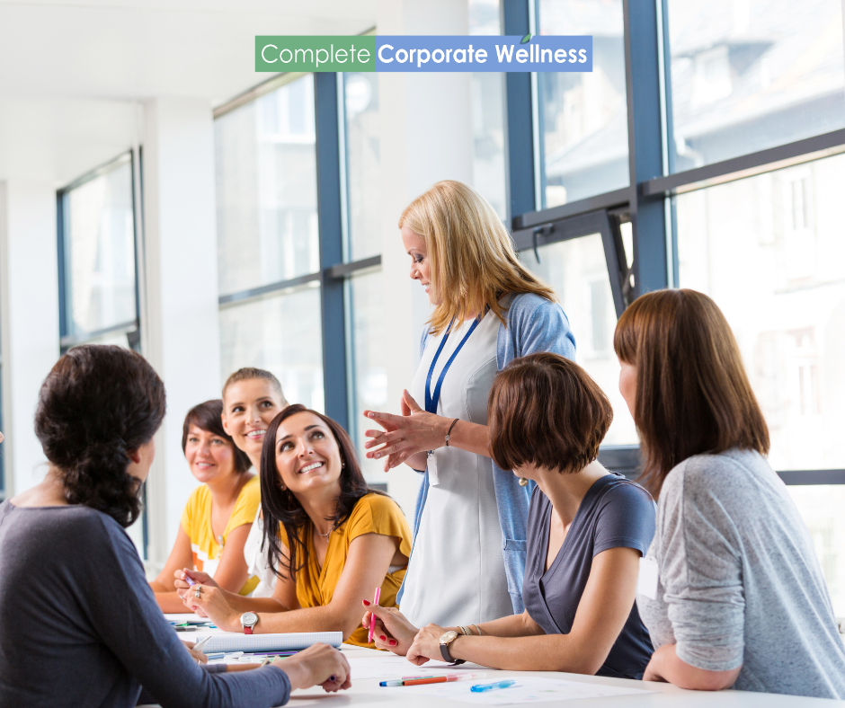 Group of women in a business meeting, one woman standing and presenting, seated women listening attentively, "Complete Corporate Wellness" sign above.