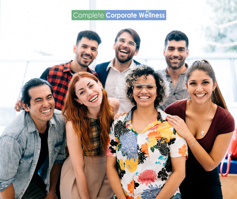 A diverse group of seven smiling people stands together in an office environment. A sign reads 'Complete Corporate Wellness' above them.