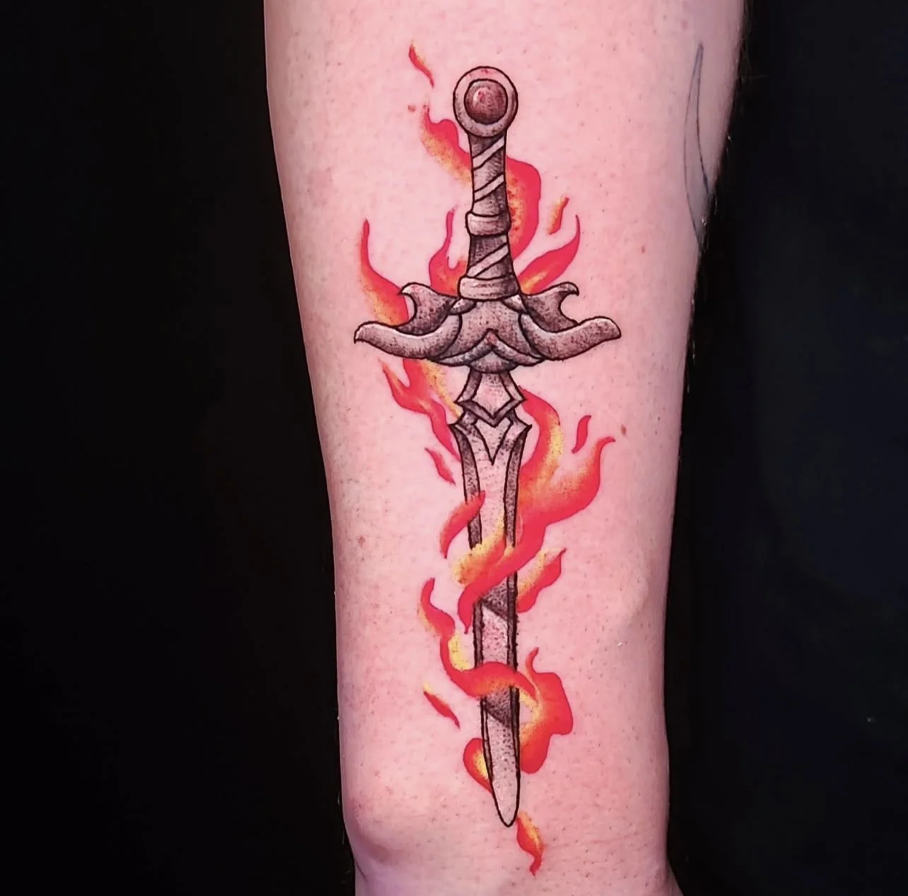 Tattoo of a sword with a detailed hilt and blade, surrounded by red and orange flames.