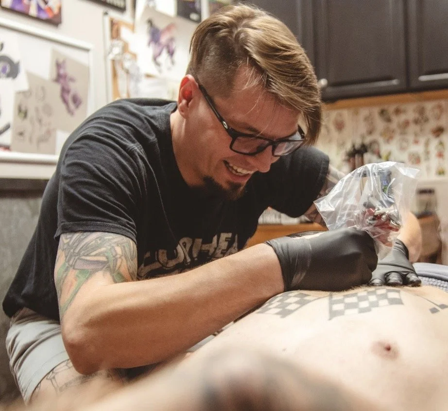 Man with tattoos, wearing glasses, a backwards cap, and a light-colored T-shirt, is sitting at a table being tattooed by an artist with a tattooed arm wearing a black glove.