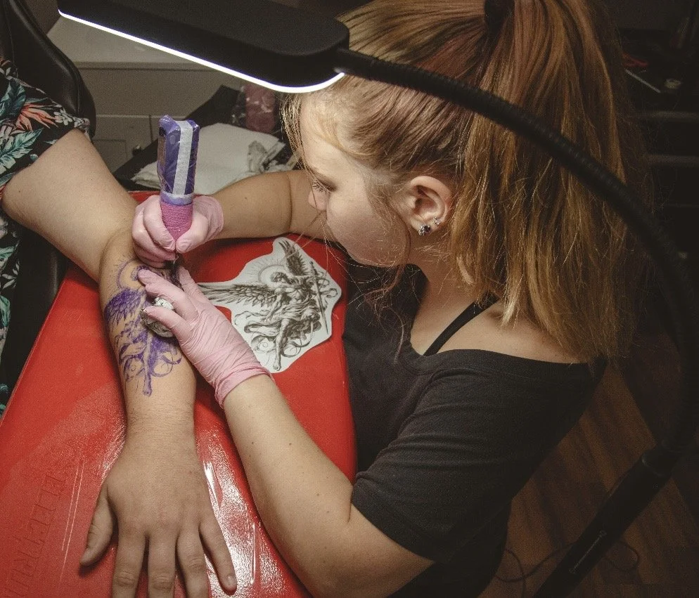 A woman with short blond hair getting tattooed on her forearm by a tattoo artist in a black shirt and gloves, in a tattoo studio.