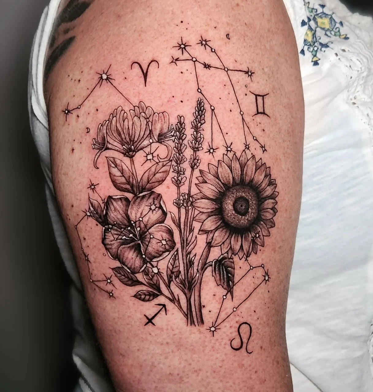 Tattoo of flowers and zodiac constellations on person's arm, including sunflower, lavender, and other flowers, with stars and zodiac symbols for Aries, Virgo, Gemini, and Leo.