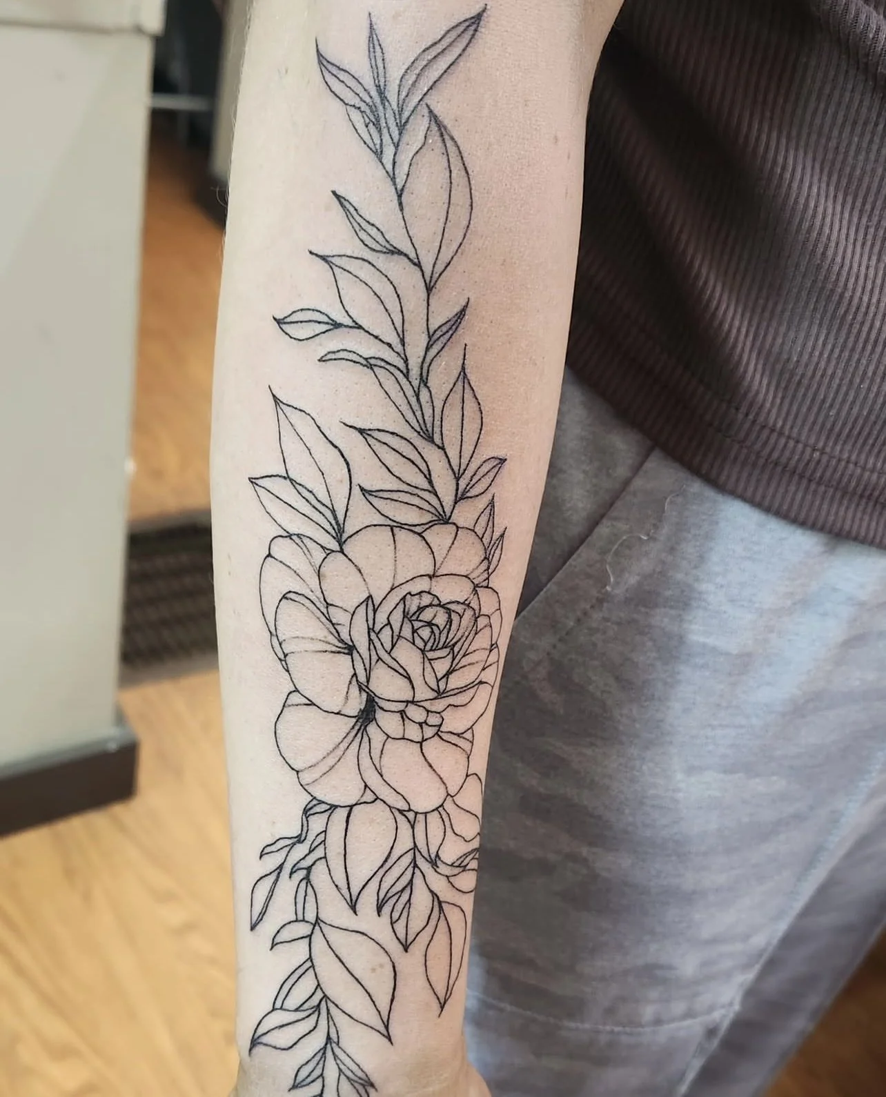 A line art tattoo of a large blooming rose with detailed leaves running down the person's forearm.