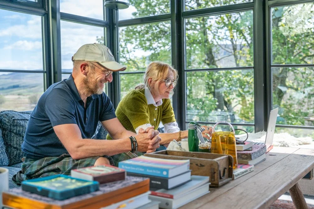 Two people, a man and a woman, sitting at a large wooden table in a bright room with floor-to-ceiling windows, surrounded by books, a laptop, and drinks, enjoying a conversation or work.