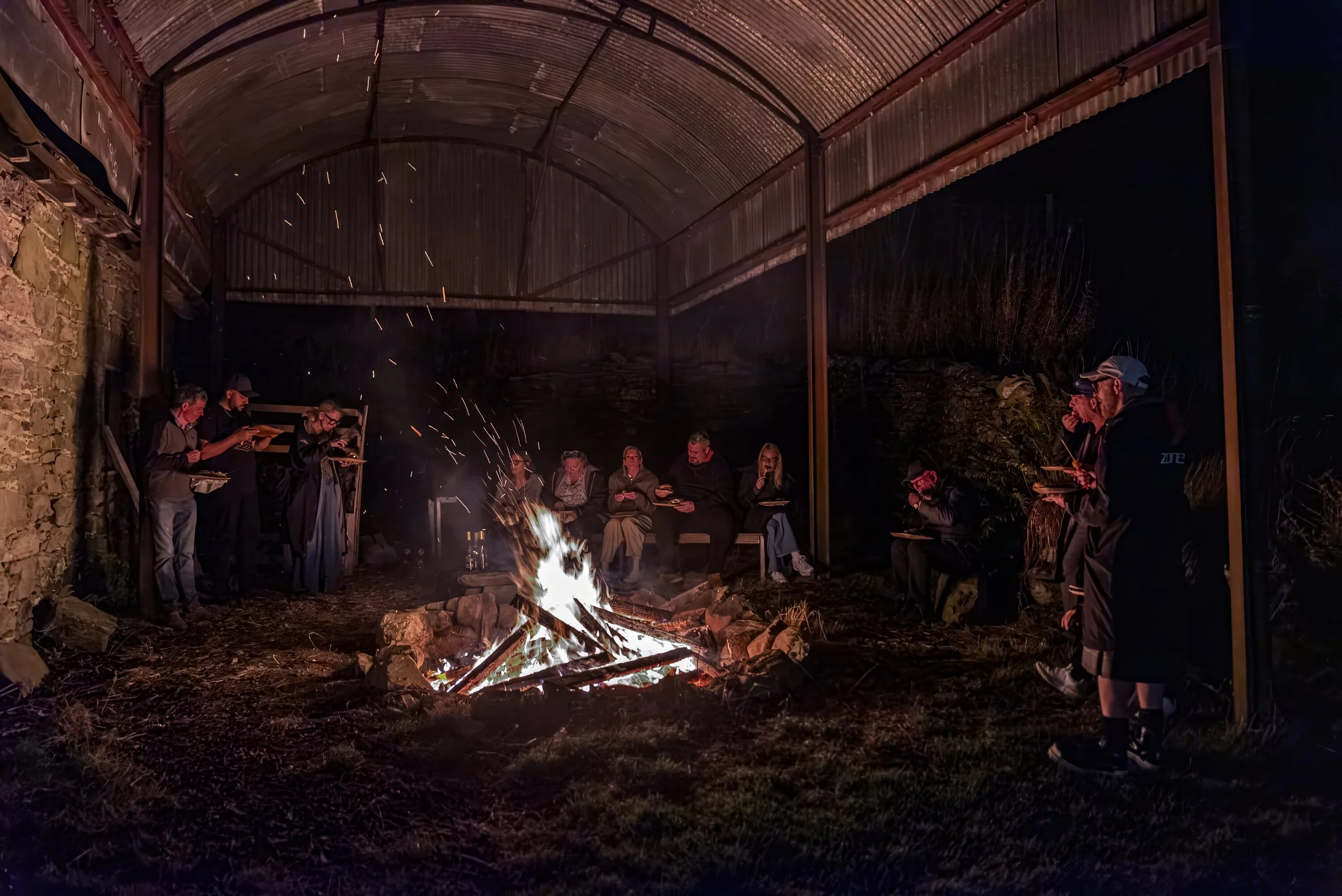 Group of people sitting and standing around a campfire under a large metal shelter at night.
