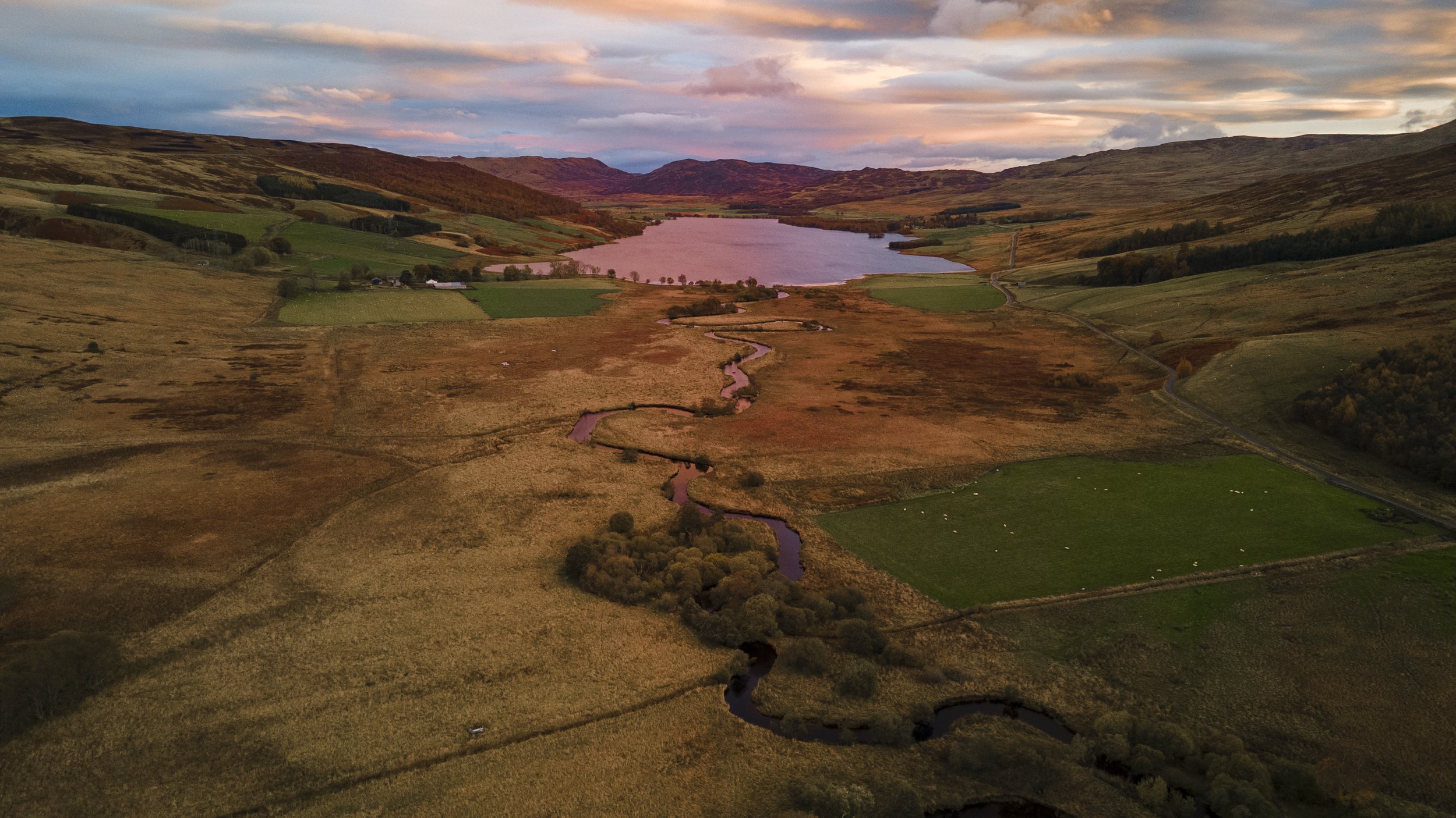 Aerial view of a rural landscape with a winding river, patches of green fields, a lake in the distance, and rolling hills during sunset.