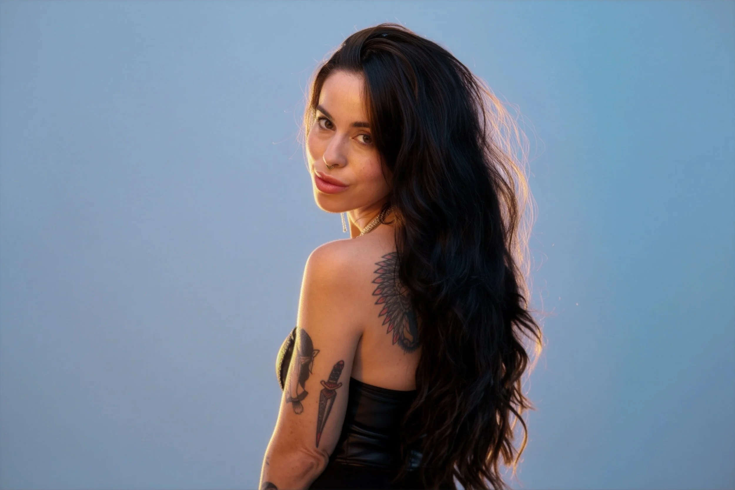 A woman with long black wavy hair, wearing a sleeveless brown top, tattoos on her arms, and jewelry including bracelets, necklaces, and rings, stands against a plain white background. She is winking and touching her neck with one hand.