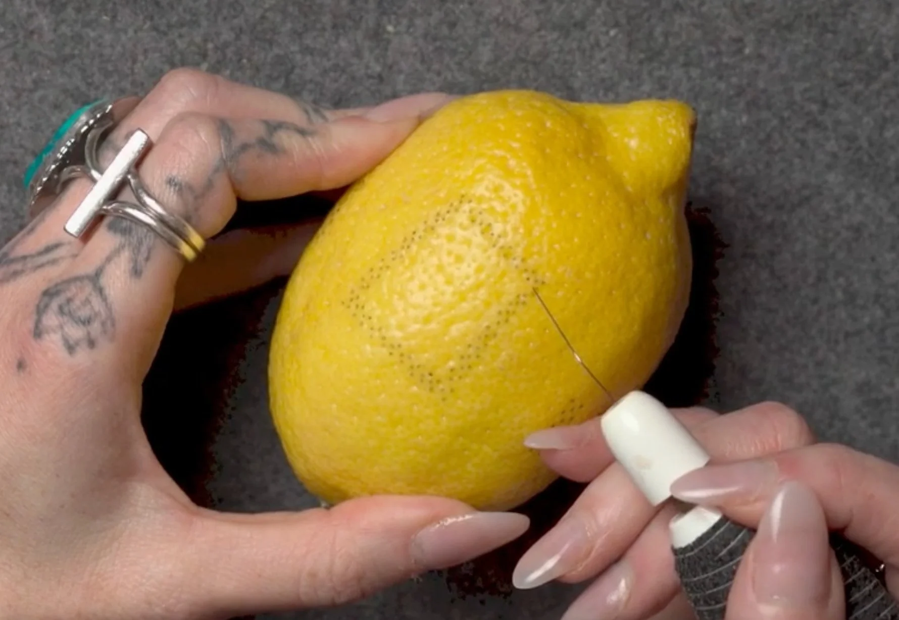 Person is threading a needle through a yellow lemon, with hands adorned with tattoos and rings, on a dark surface.