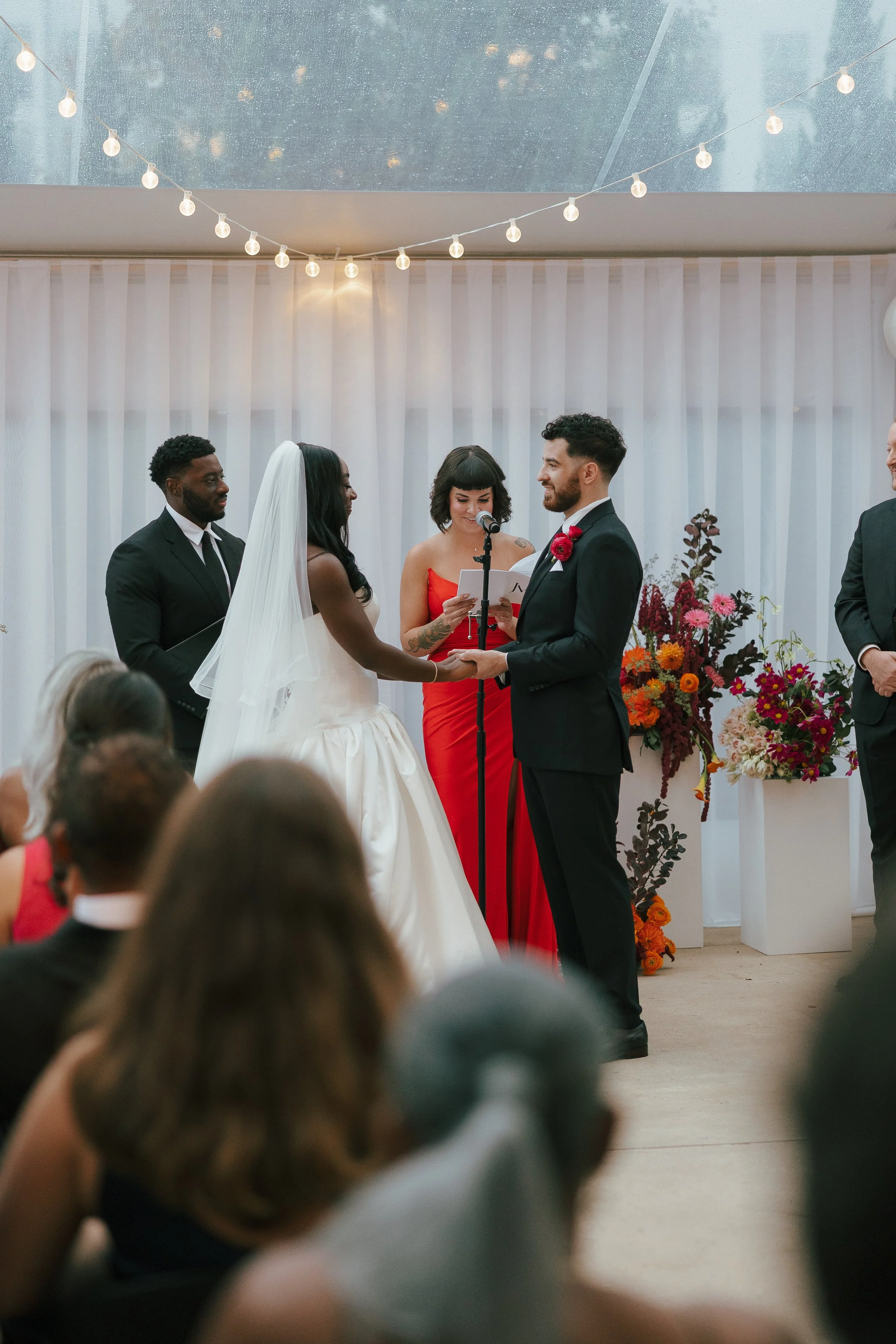 A wedding ceremony with a couple exchanging vows, surrounded by officiant, witnesses, and floral arrangements, indoors with string lights and white curtains.