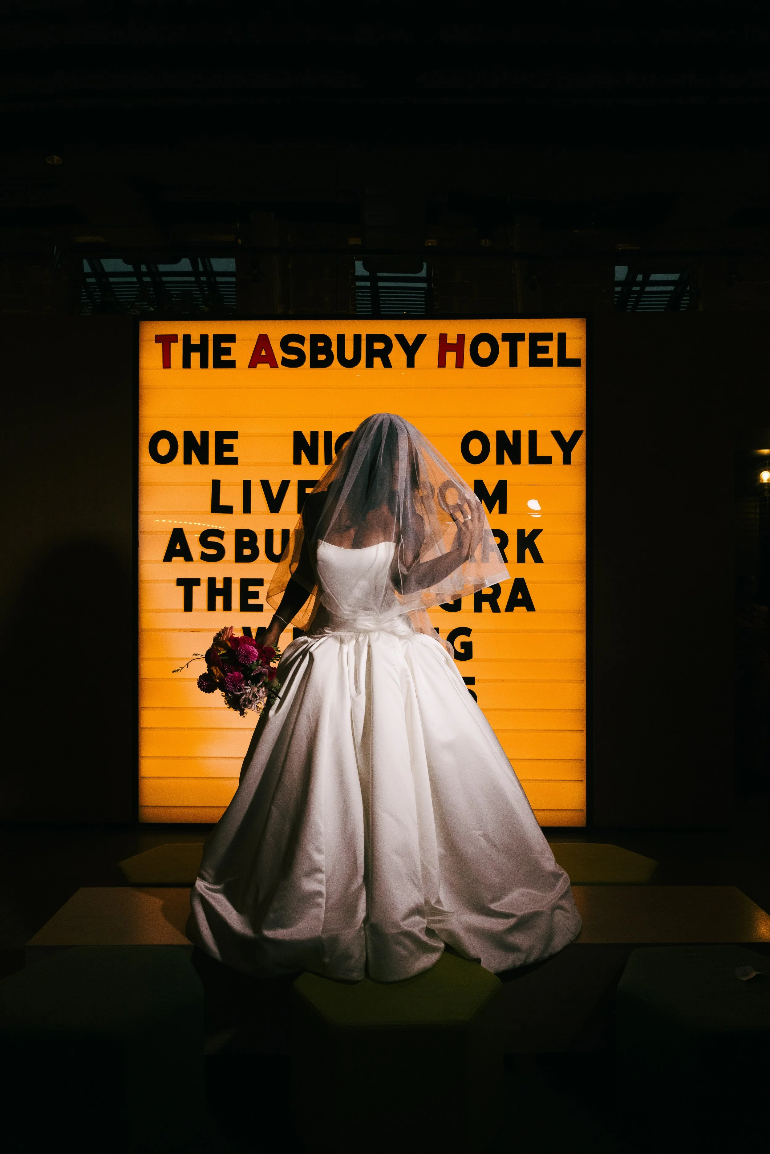 A woman in a white wedding gown with a veil holds a bouquet of flowers, standing in front of an illuminated sign with black and red text.
