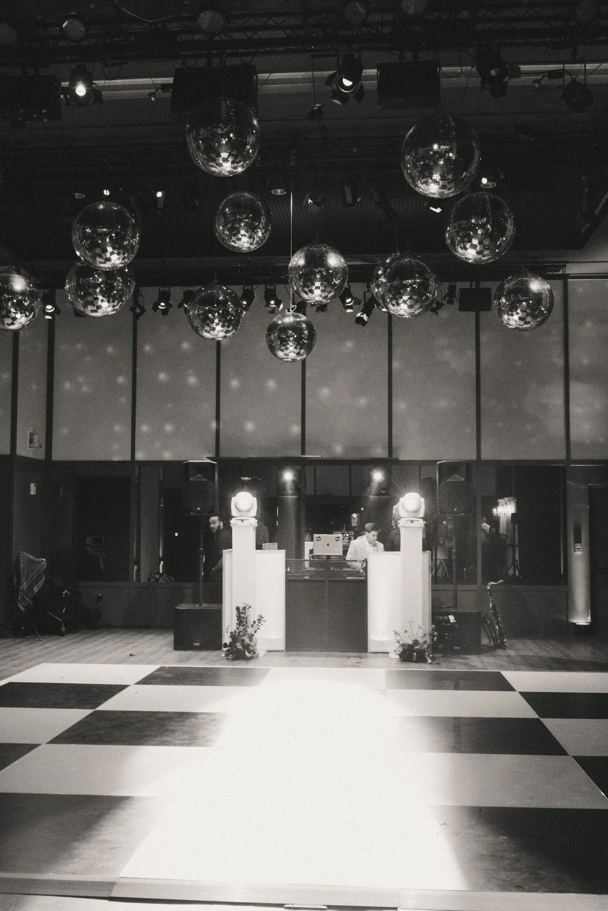 Empty dance floor with a black and white checkered pattern, ceiling decorated with multiple disco balls, and a DJ booth with two DJs and large speakers in the background.