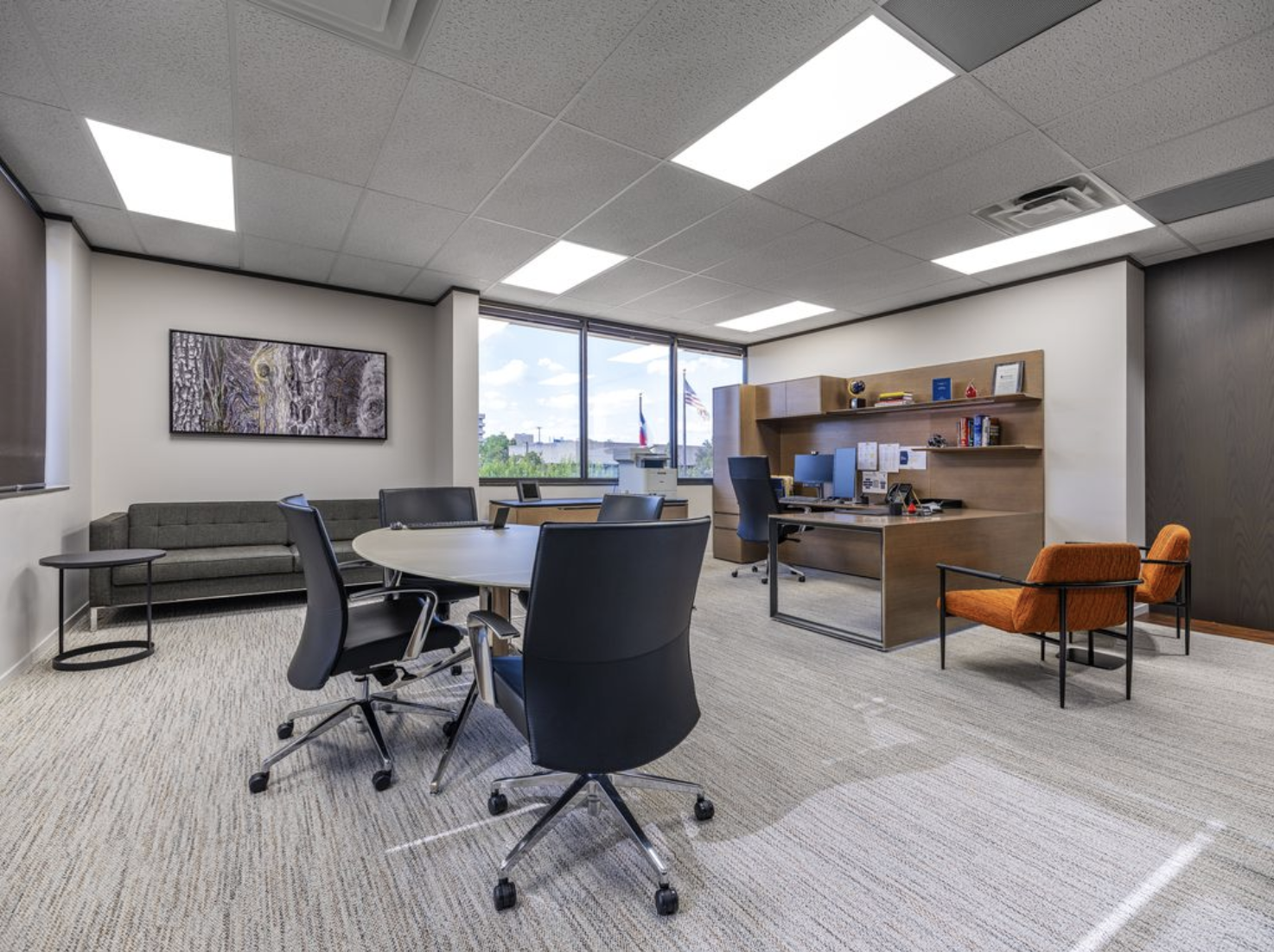 An office room with a round table and four black office chairs, a gray sofa against the wall, a large window with a view outside, a wooden desk with computer monitors and office supplies, and orange chairs. There are ceiling lights and a abstract wal