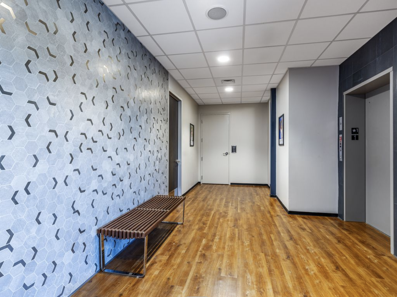 Interior of a modern building hallway with hardwood floor, a bench on the left, decorative wall on the left, and an elevator to the right. Two framed pictures on the walls, ceiling with recessed lighting, and a closed door at the end.