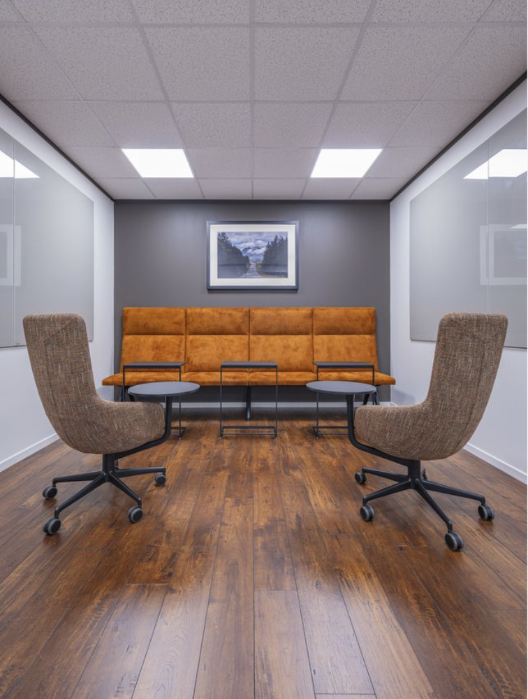 An empty office meeting room with two beige swivel chairs facing a tan upholstered bench and a glass-top table, a framed landscape photograph on a gray wall, and wood flooring.