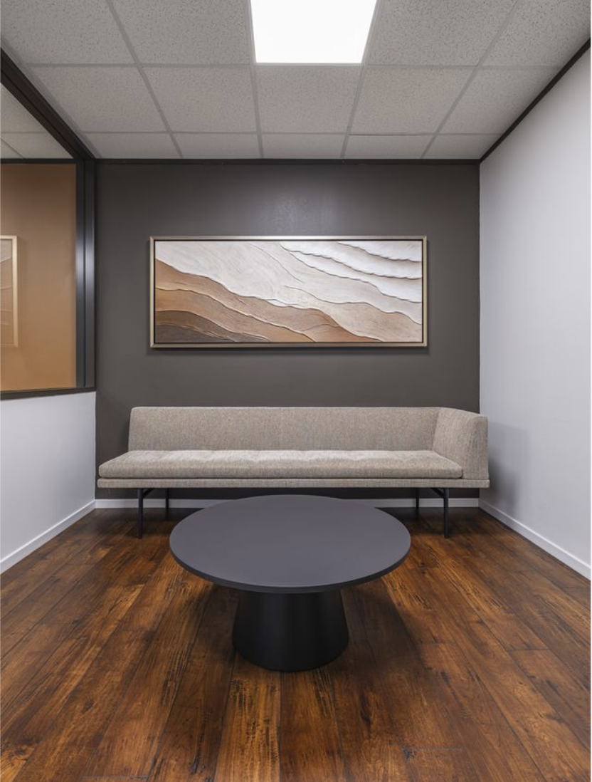 An office waiting area with a beige sofa against a dark gray wall, a round black coffee table in front, a framed abstract artwork above, and a ceiling with overhead fluorescent lighting.