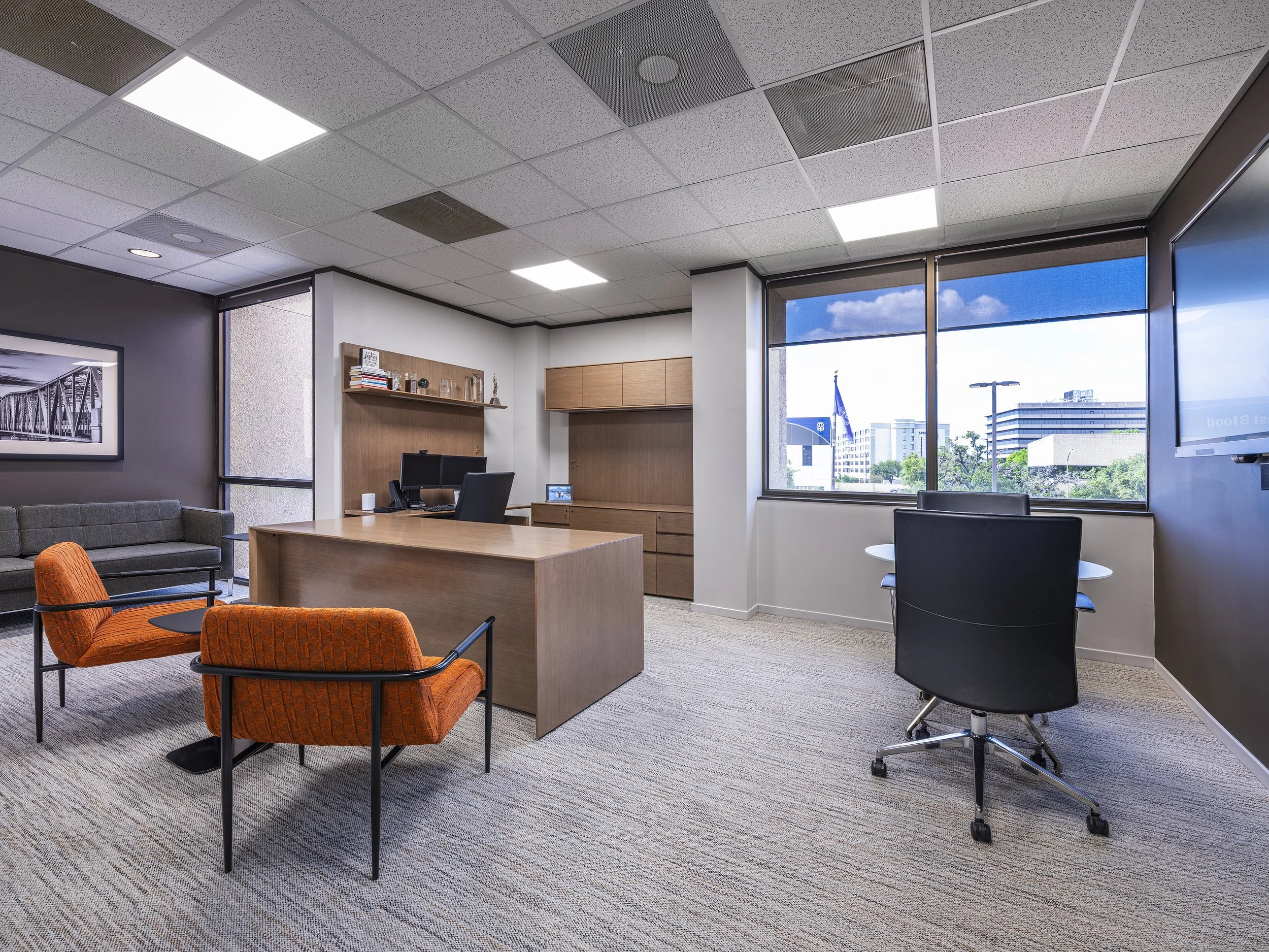 An office with large windows showing a cityscape with buildings and trees outside. Inside, there is a wooden desk with a computer monitor, a small framed photo, and cabinets behind it. Seating includes a grey sofa, orange chairs, and black office cha