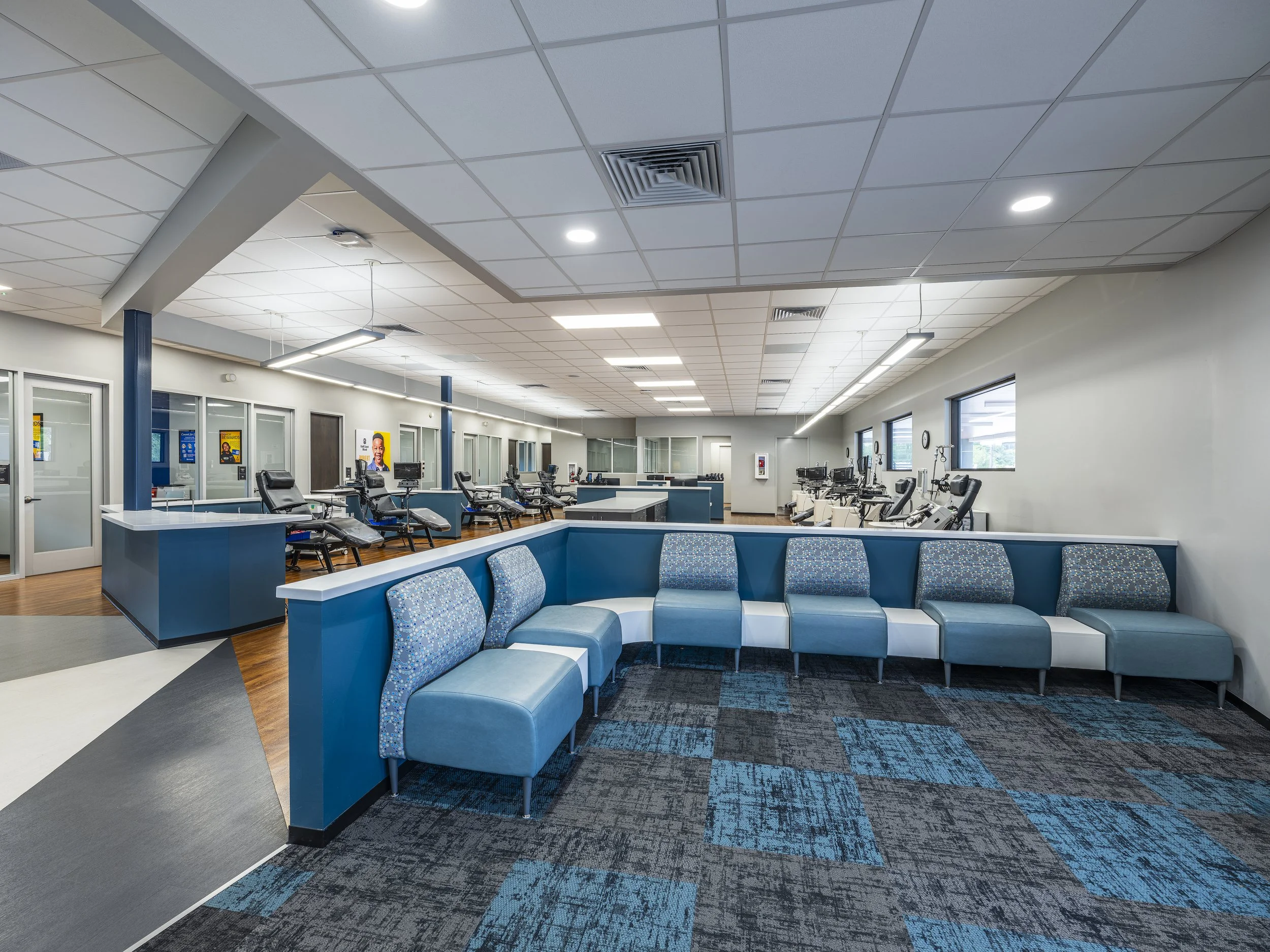 The interior of a modern medical or clinic waiting room with blue chairs arranged in a semi-circle, reception counters, and examination rooms in the background.