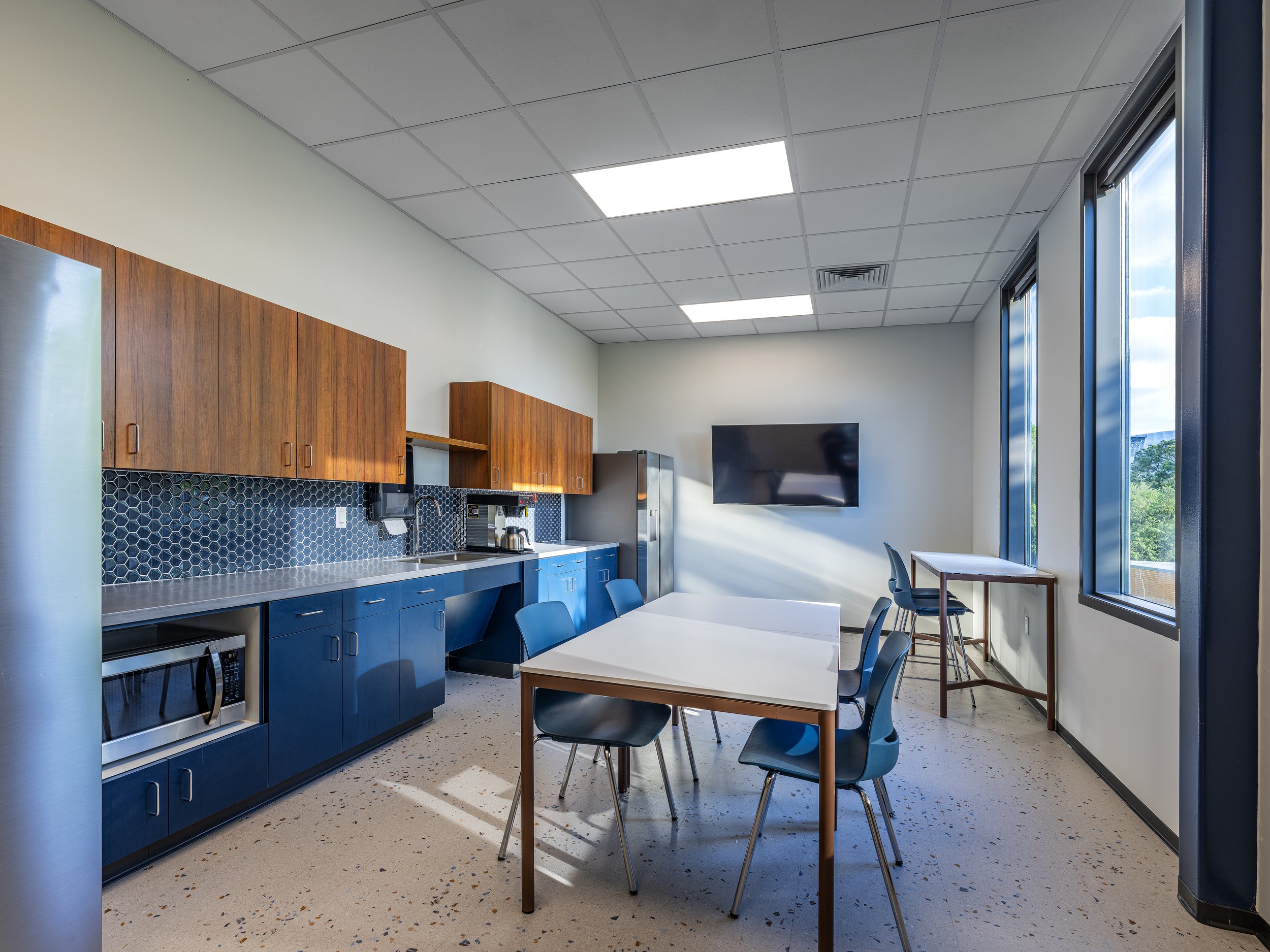 A kitchen with blue cabinets, wooden wall cabinets, a black backsplash with honeycomb tiles, and a white countertop, with a window showing trees outside.
