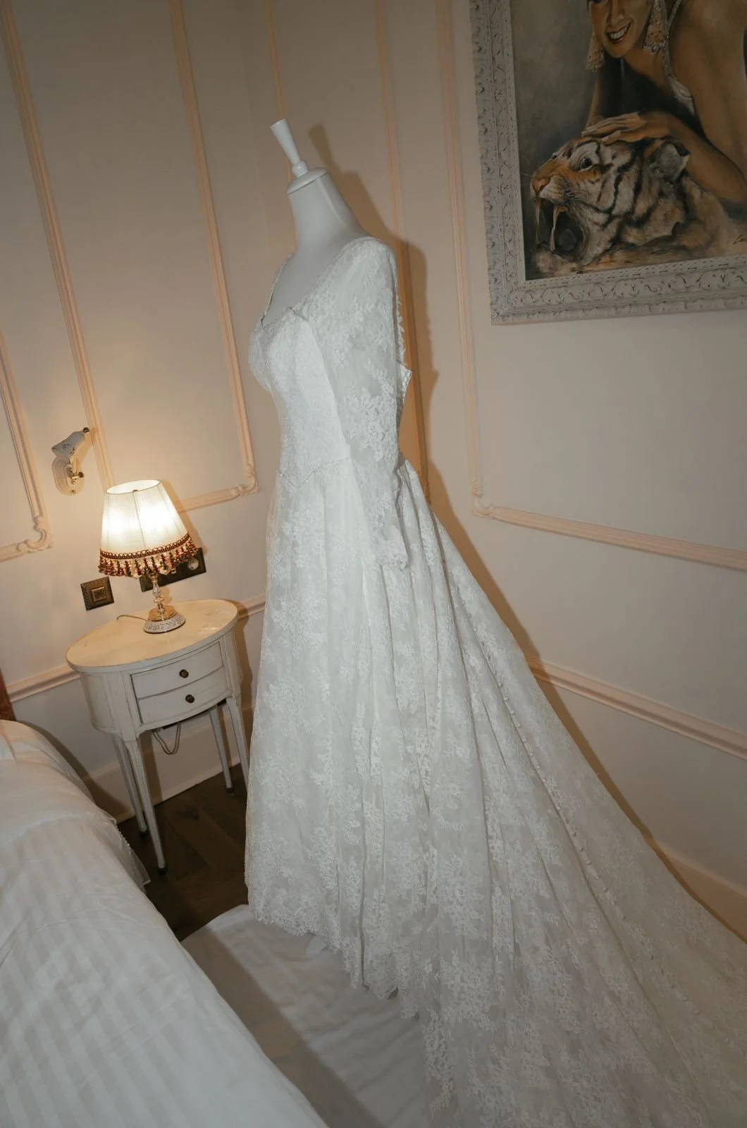A white lace wedding dress displayed on a mannequin in a room with beige walls, a small white side table with a lamp, and a framed painting of a woman and a tiger.