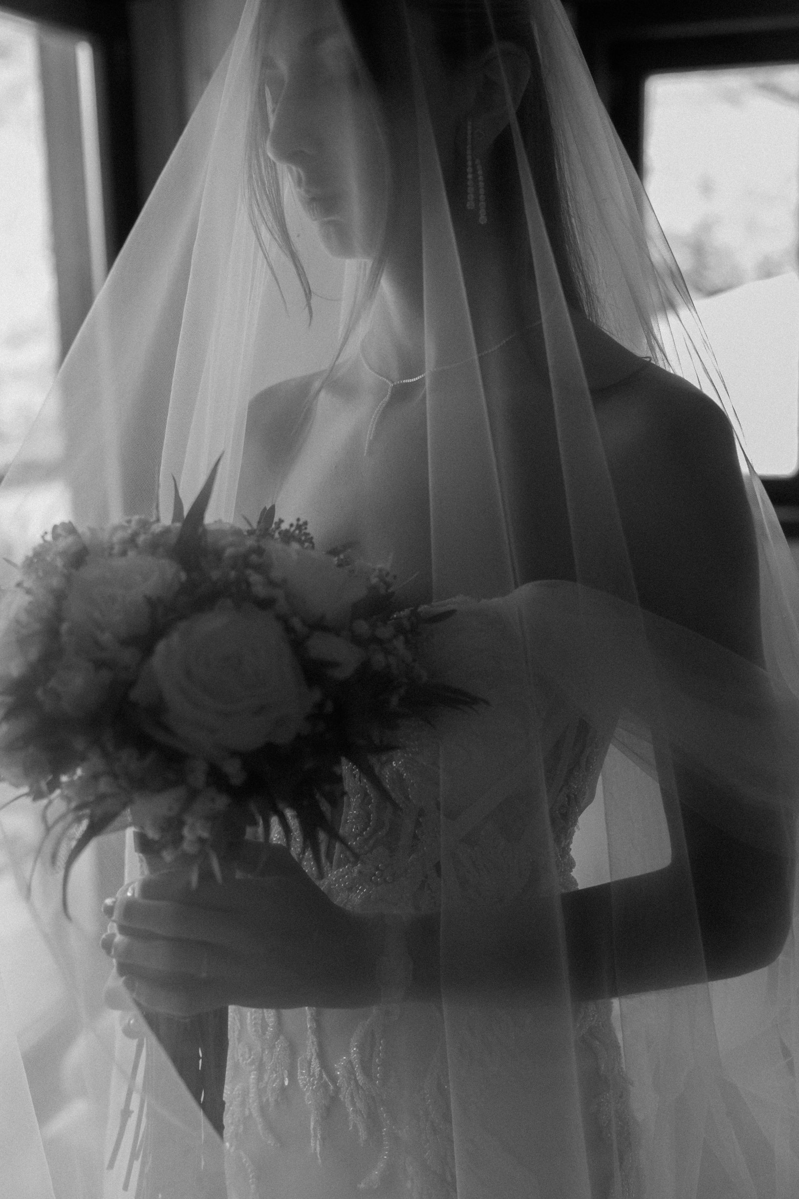 A black and white photo of a bride holding a bouquet of flowers, wearing a wedding dress and veil, with her face partially visible as she looks down.