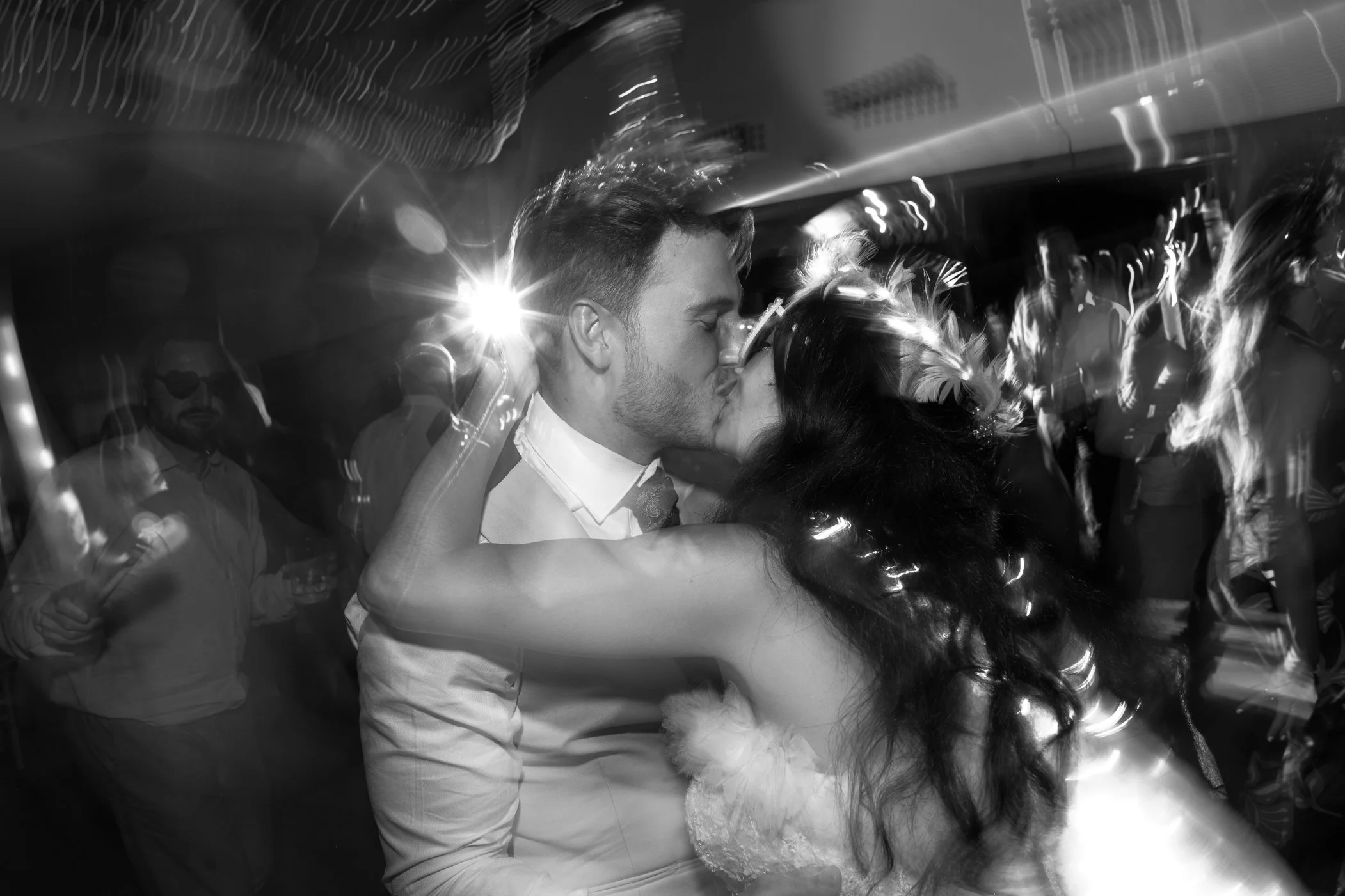 A couple sharing a kiss at a wedding reception, with the groom in a dress shirt and tie and the bride in a wedding dress and veil, surrounded by guests and blurred lights.