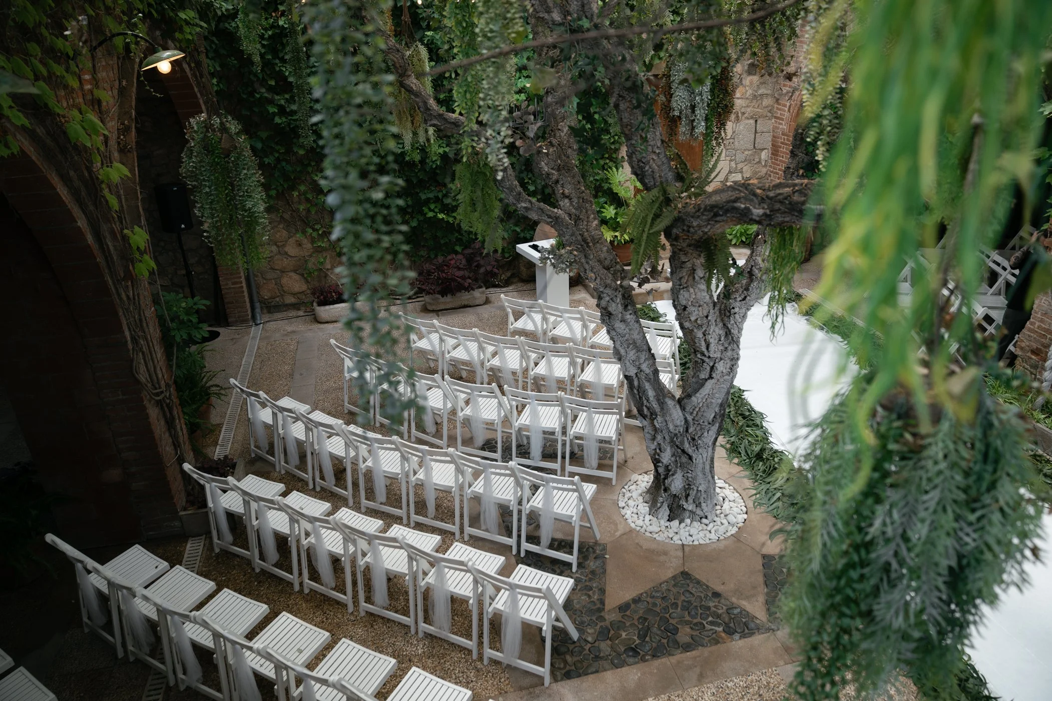 Outdoor wedding ceremony setup with white chairs arranged in rows facing an altar, under a large tree with hanging greenery, surrounded by stone and brick walls.