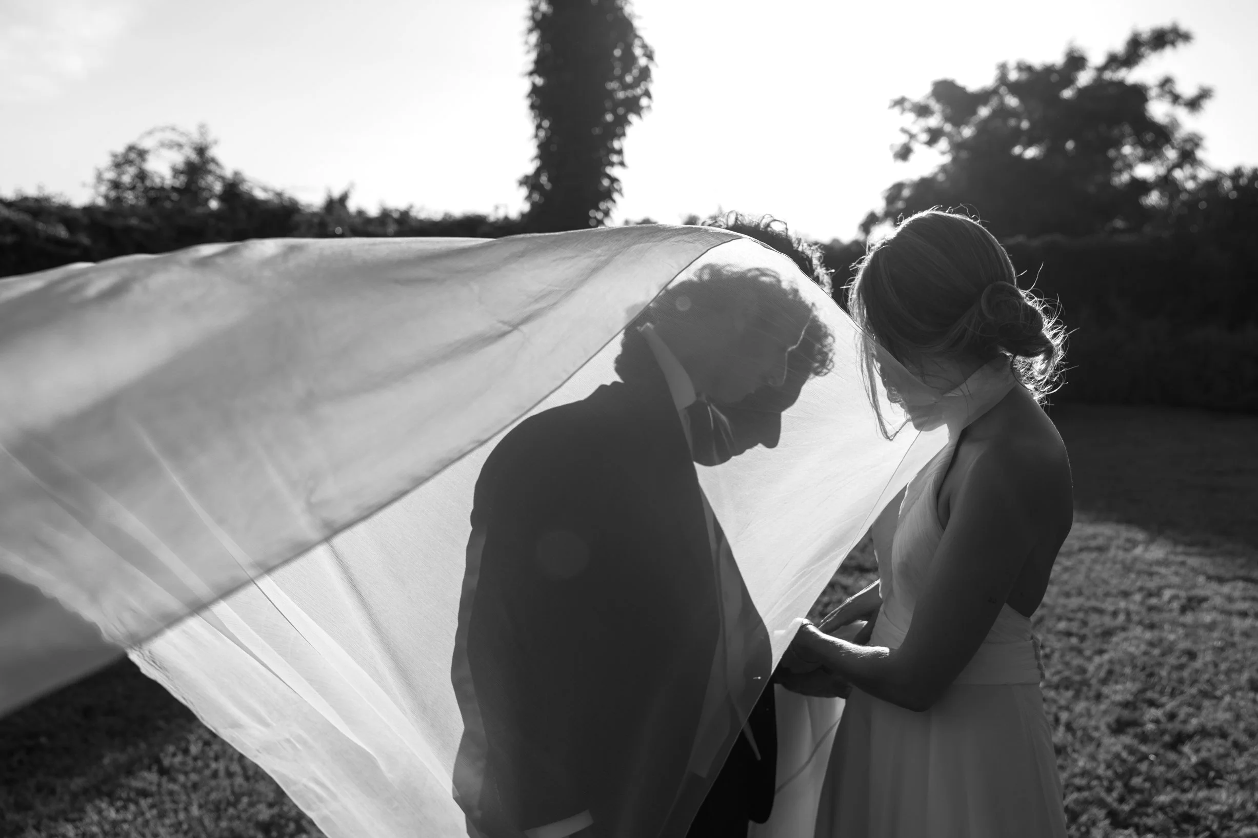 A black and white photo of a bride and groom under a wedding veil outdoors at sunset, holding hands and facing each other.