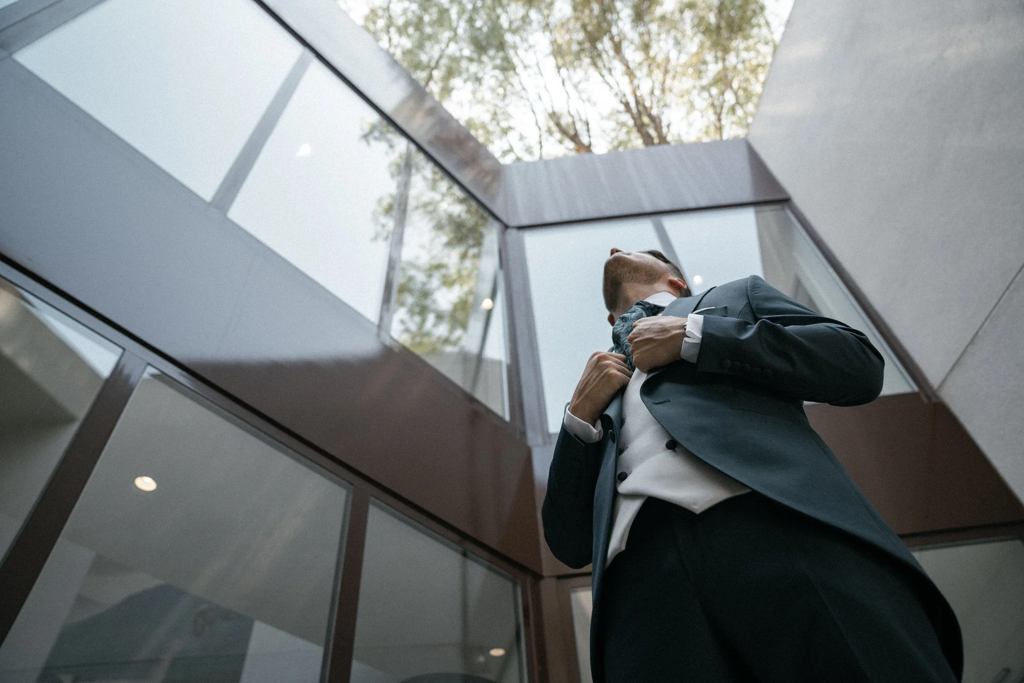A man in a suit adjusting his jacket outside a modern building, viewed from below.