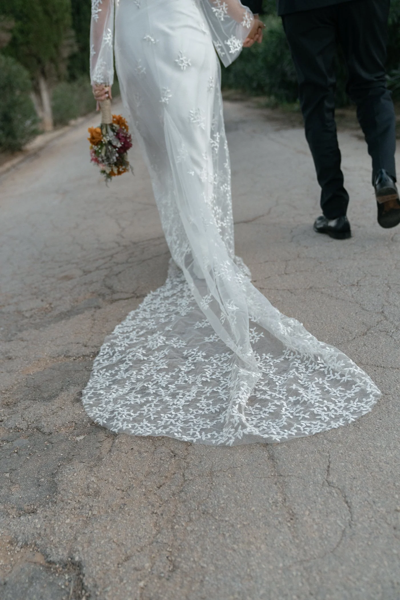 Bride in a wedding dress with a long lace train, holding a bouquet, walking on an outdoor cracked pavement, with a groom walking beside her.