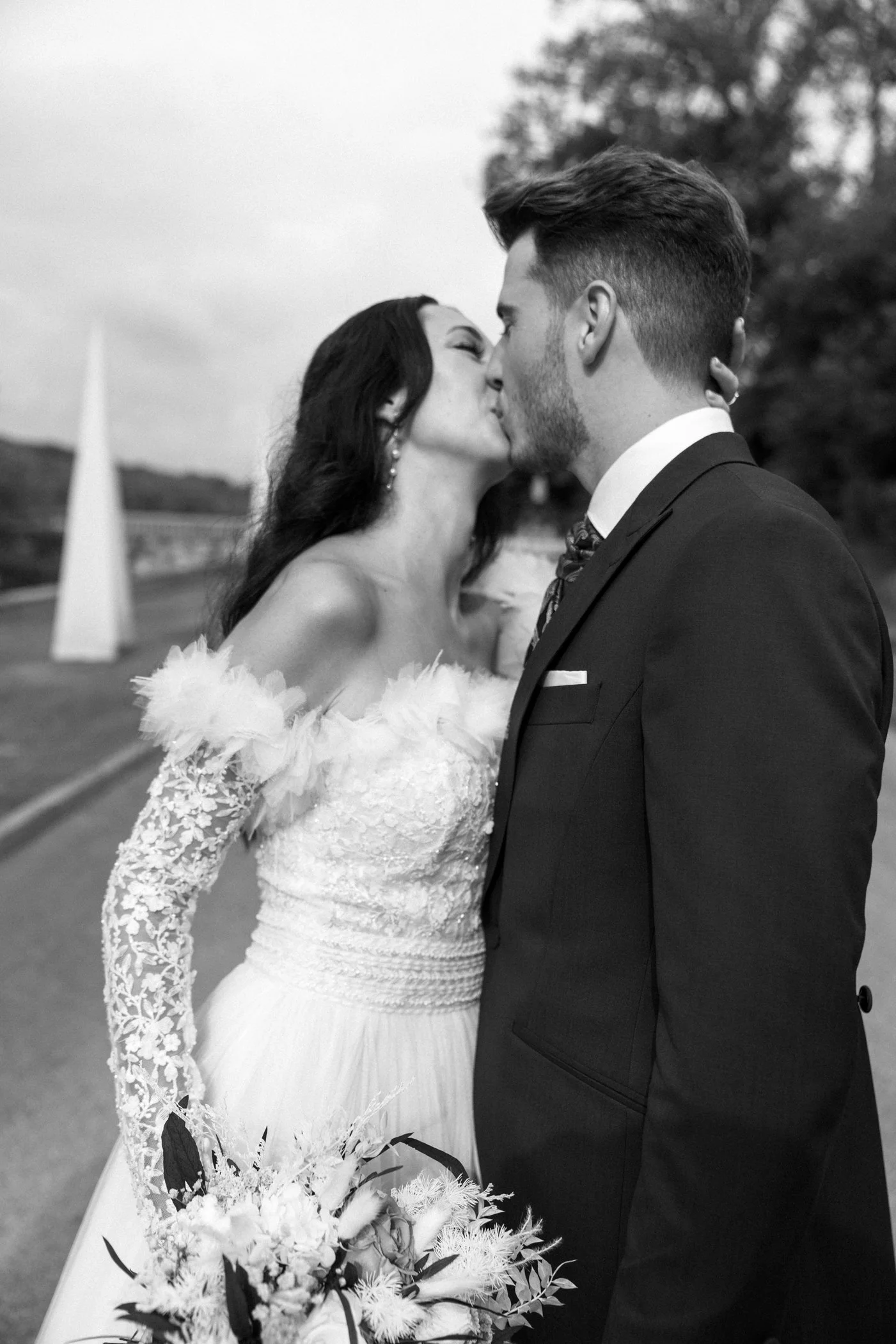 A black and white photo of a bride and groom sharing a kiss outdoors, with the bride holding a bouquet of flowers.