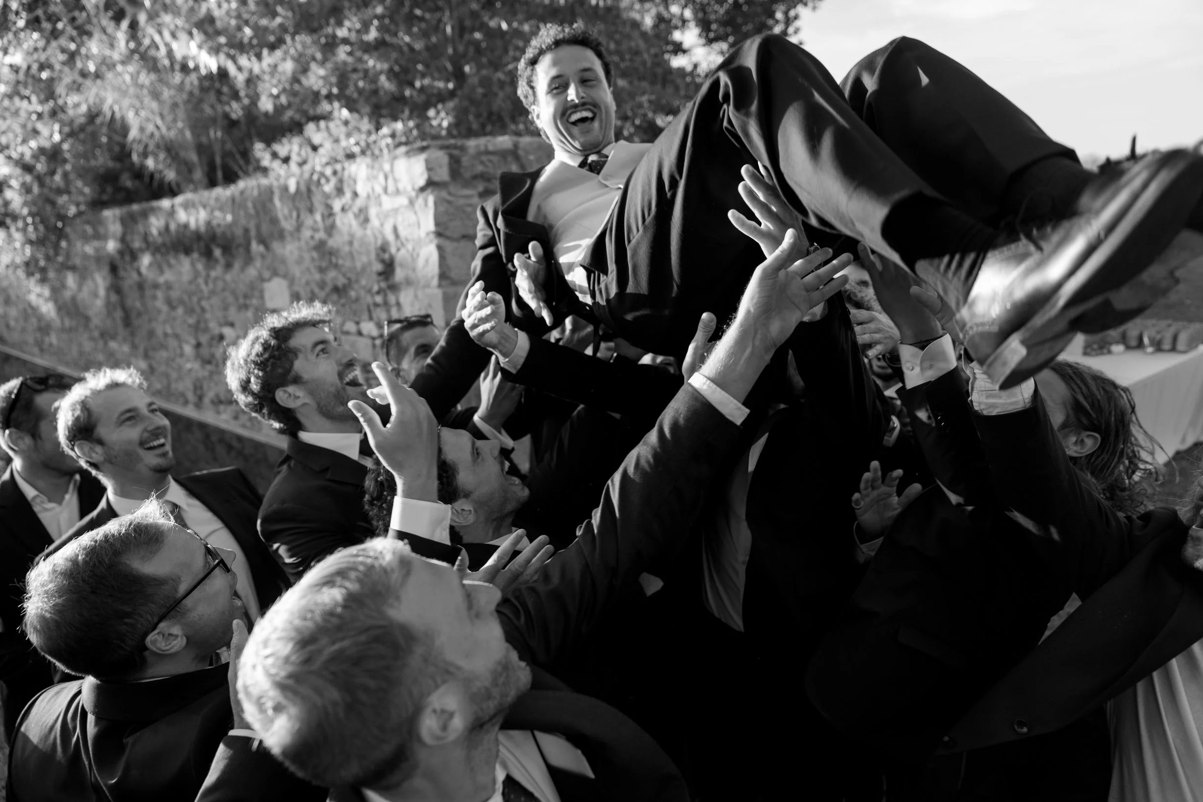 A group of men in suits lifting a smiling man in a tuxedo into the air outdoors during a celebration.