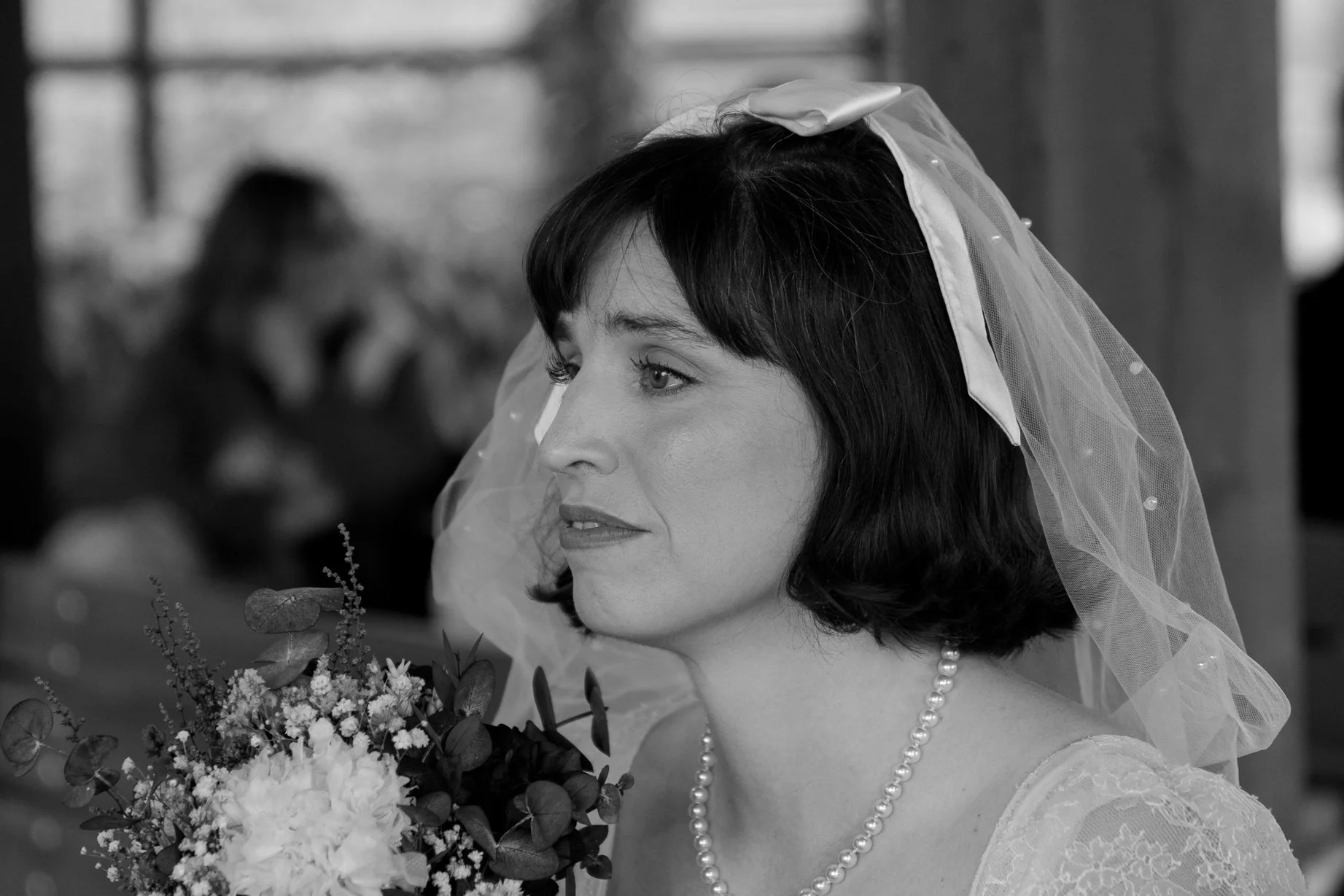 A woman with dark short hair and light eyes, dressed in a wedding gown with pearls, wearing a veil, holding a bouquet of flowers, during a wedding ceremony.