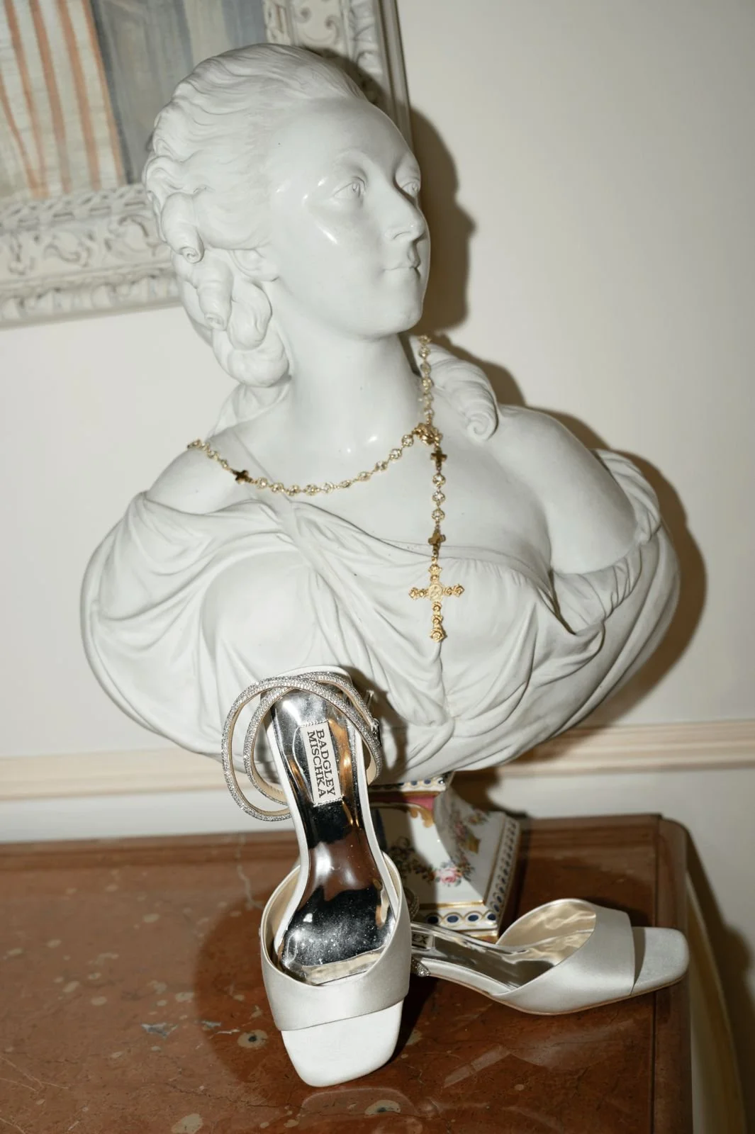 A white marble bust of a woman wearing a gold necklace and holding a pair of silver high-heeled shoes marked 'Badgley Mischka'. The bust is placed on a wooden surface against a neutral wall.
