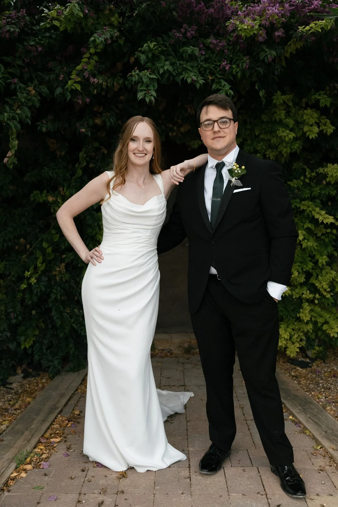 A bride and groom pose together outdoors in front of lush green foliage. The bride wears a white wedding gown and the groom a black suit with a tie and boutonniere.