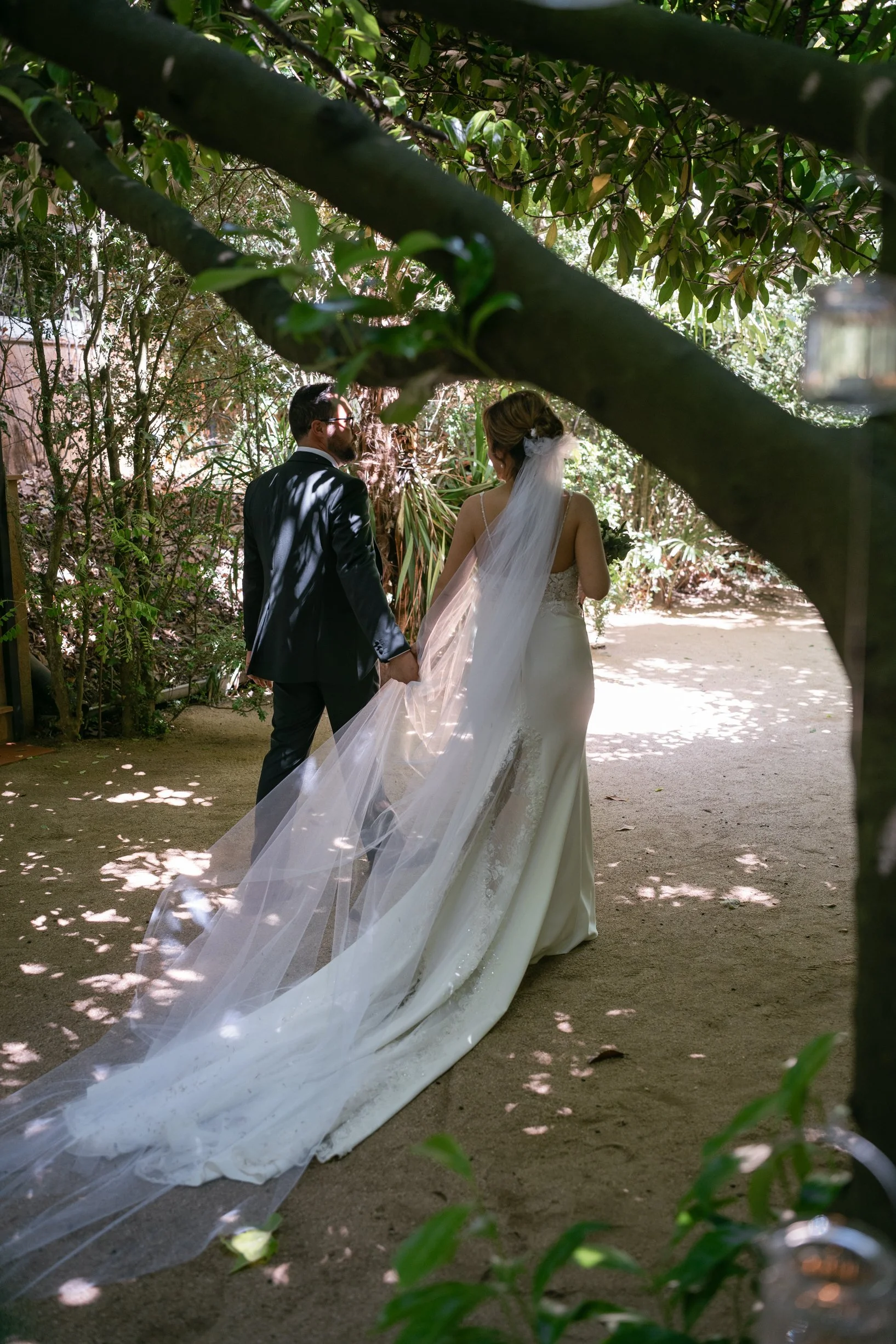 A bride and groom walking hand in hand through a shaded outdoor garden, with the bride wearing a long white wedding dress and veil, and the groom in a black suit.
