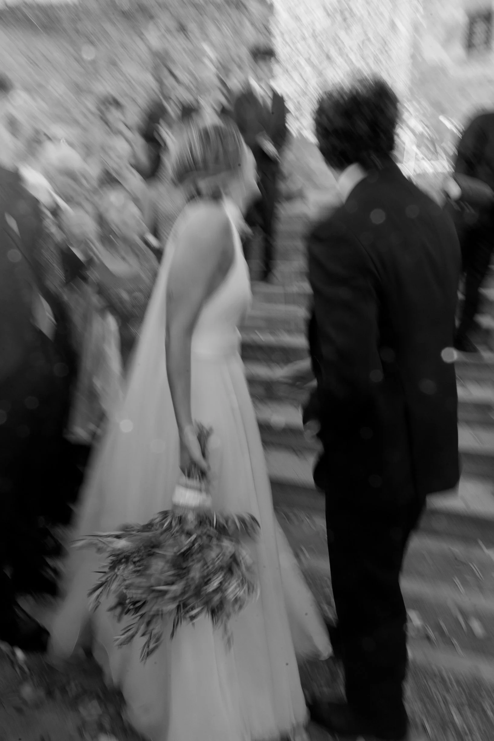 A black-and-white photo of a bride and groom holding hands during a wedding ceremony, with guests in the background.