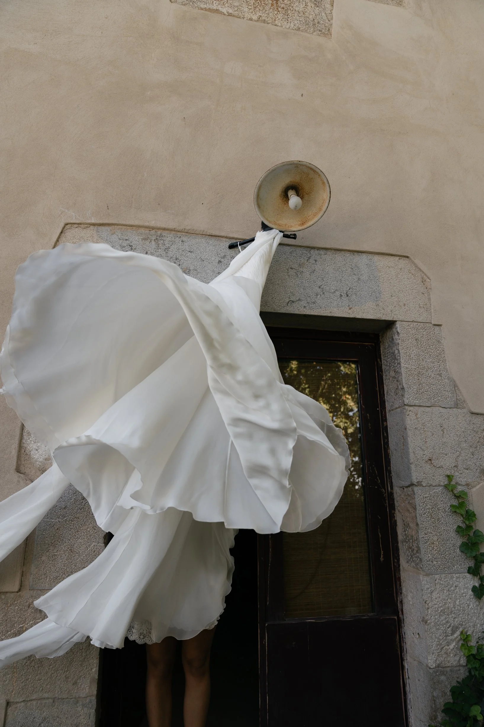A person wearing a white dress is standing outside a stone building. The photo is taken from below, showing the person's head and lower legs, along with a lamp and a window on the building.
